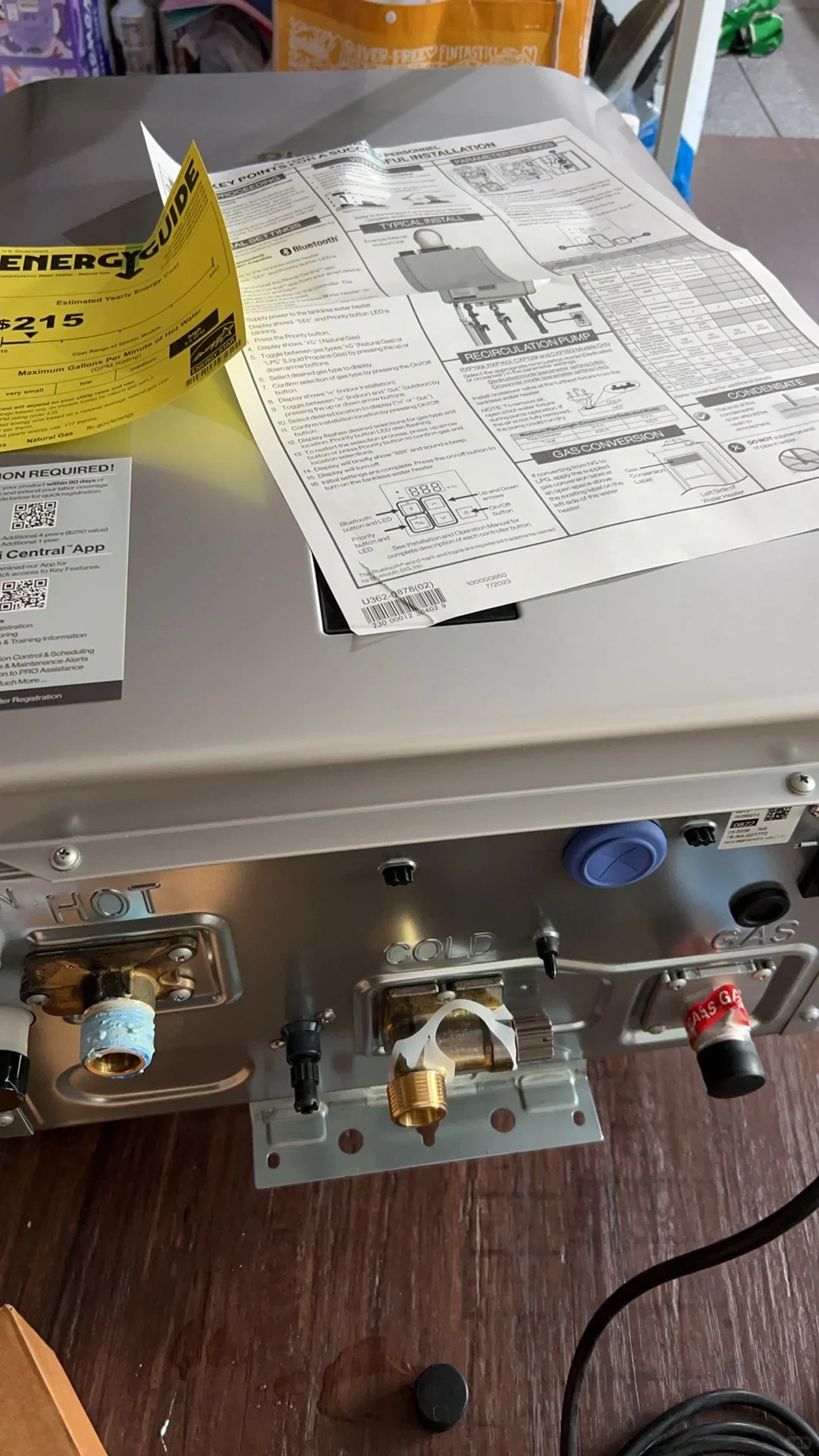 The underside of the Rinnai water heater shows connections marked HOT, COLD and GAS, with an installation sheet and EnergyGuide label lying across its casing.