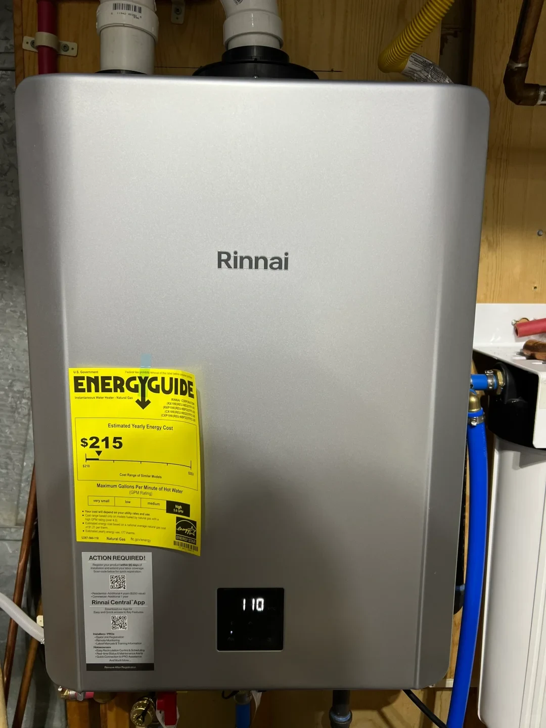 A silver Rinnai tankless water heater is mounted in a wooden utility area with pipes and hoses connected above and below. Its display reads “110,” and yellow energy and white registration labels are attached to the front.