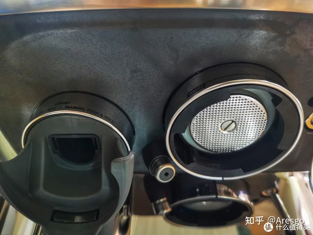 Close-up of a stainless-steel Sage espresso machine showing the grinder outlet on the left and the circular brew head with a perforated shower screen on the right.