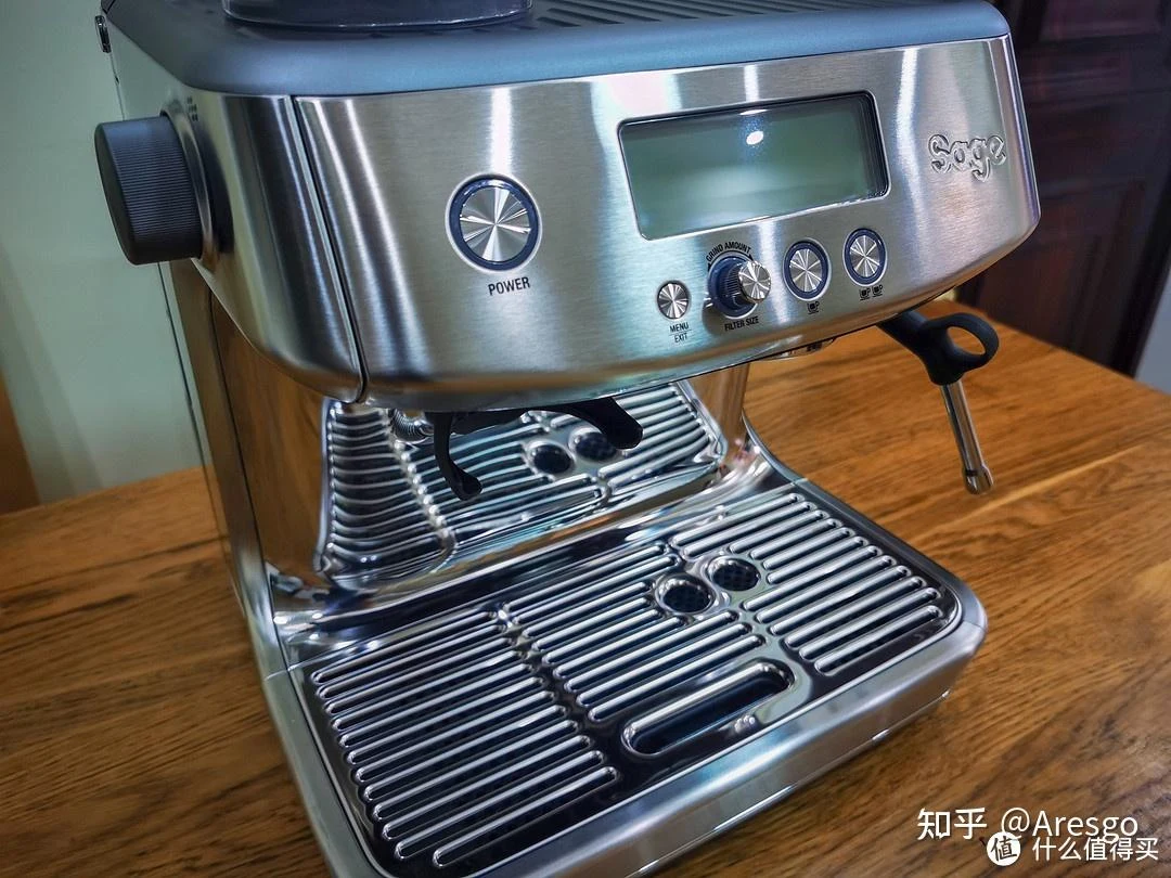 Close-up of a stainless-steel Sage espresso machine on a wooden countertop, showing its front control panel, grind-size dial, portafilter area, drip tray, bean hopper, and steam wand.