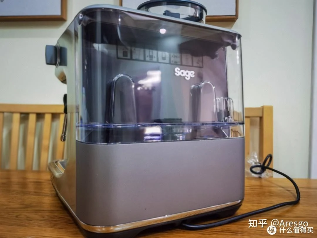 A silver Sage espresso machine with a clear upper cover sits on a wooden table in a home kitchen, with its black power cord trailing to the right.