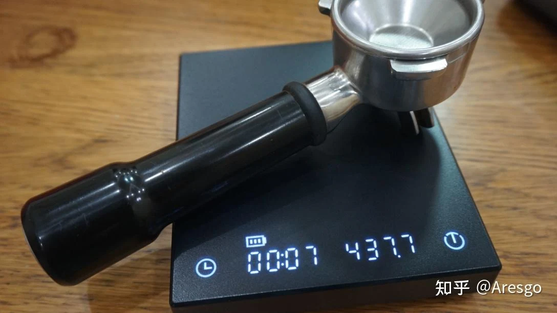 A black-handled espresso portafilter rests on a digital coffee scale on a wooden countertop; the illuminated display shows a timer of 00:07 and a reading of 43.77.