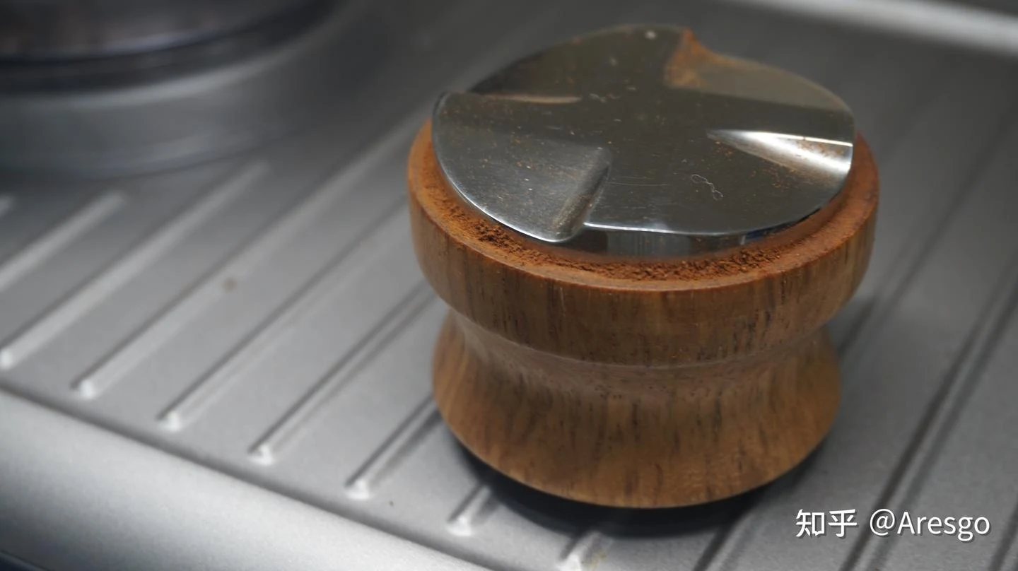 A wooden coffee distribution tool with a stainless-steel top resting on the ridged metal drip tray of an espresso machine.