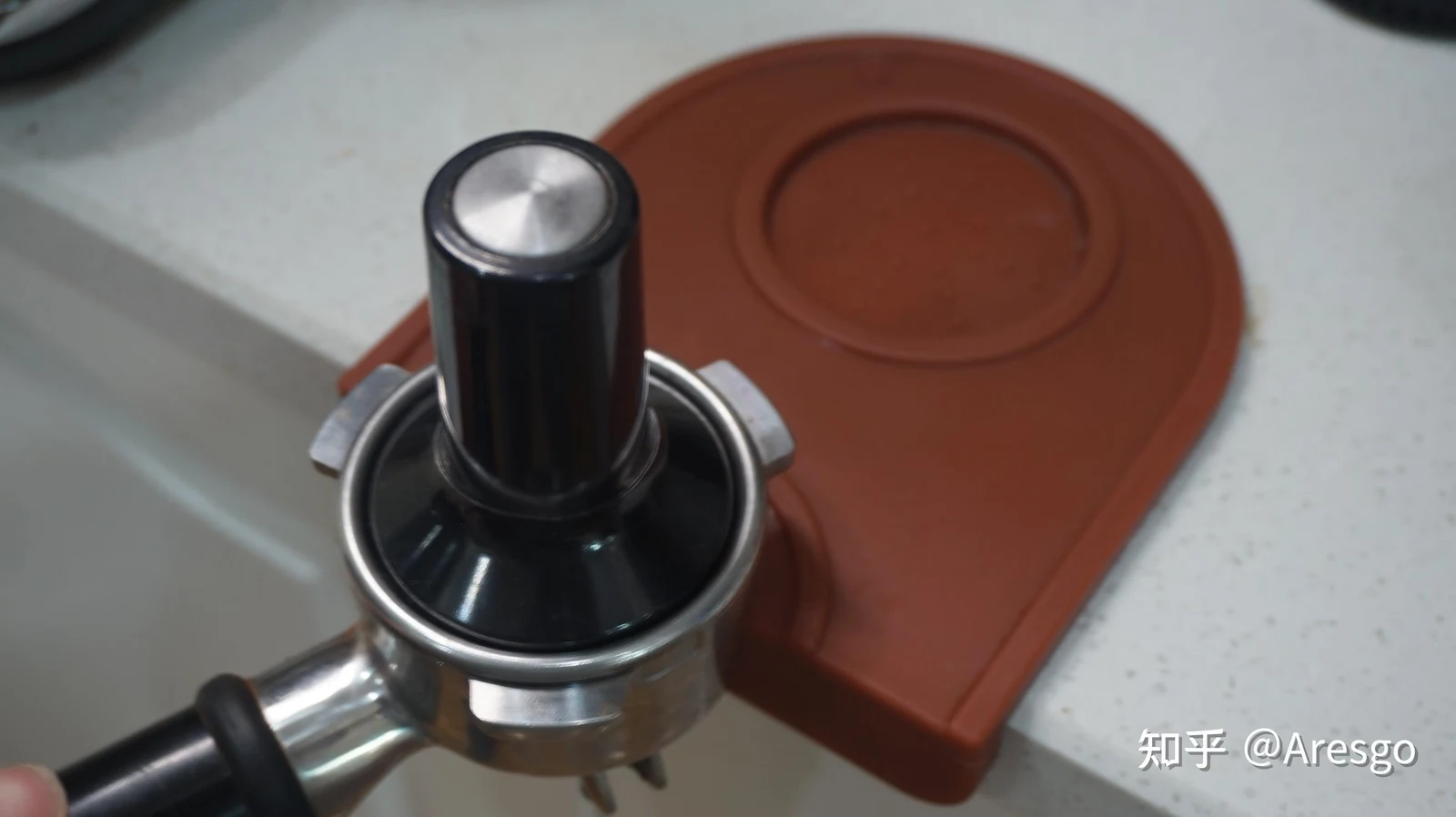 A stainless steel portafilter held over a brown silicone tamping mat on a kitchen counter edge, with a black tamper pressed into the filter basket.