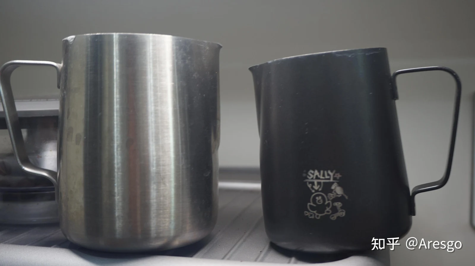 Two milk frothing pitchers side by side on a tray, featuring a stainless steel pitcher on the left and a matte black pitcher with a cartoon chick engraving on the right.