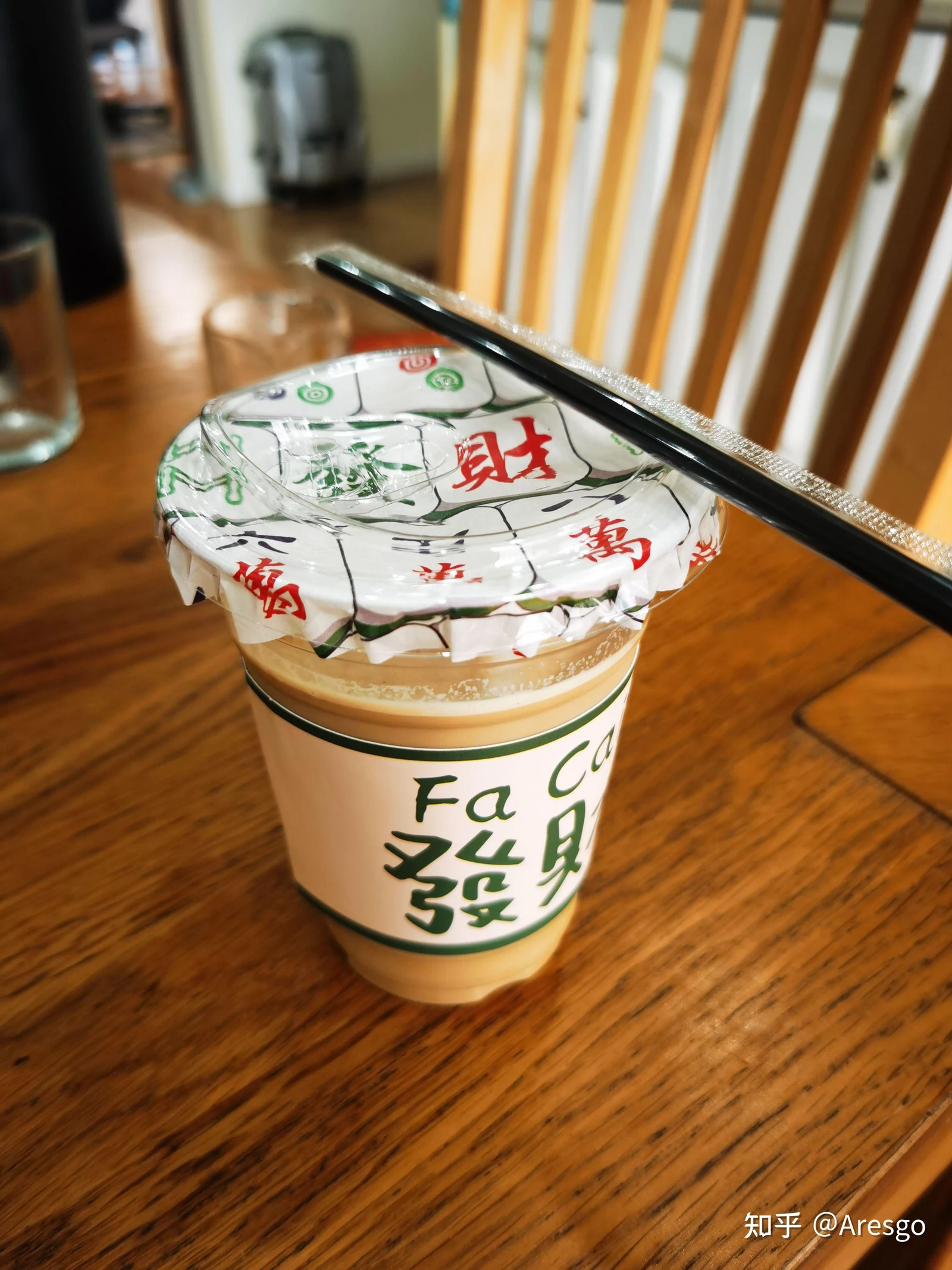 An iced coffee in a clear plastic takeaway cup sealed with mahjong-themed decorative paper and a clear lid, wrapped with a cup sleeve reading 'Fa Cai 发财', resting on a wooden table with a black straw across the top.