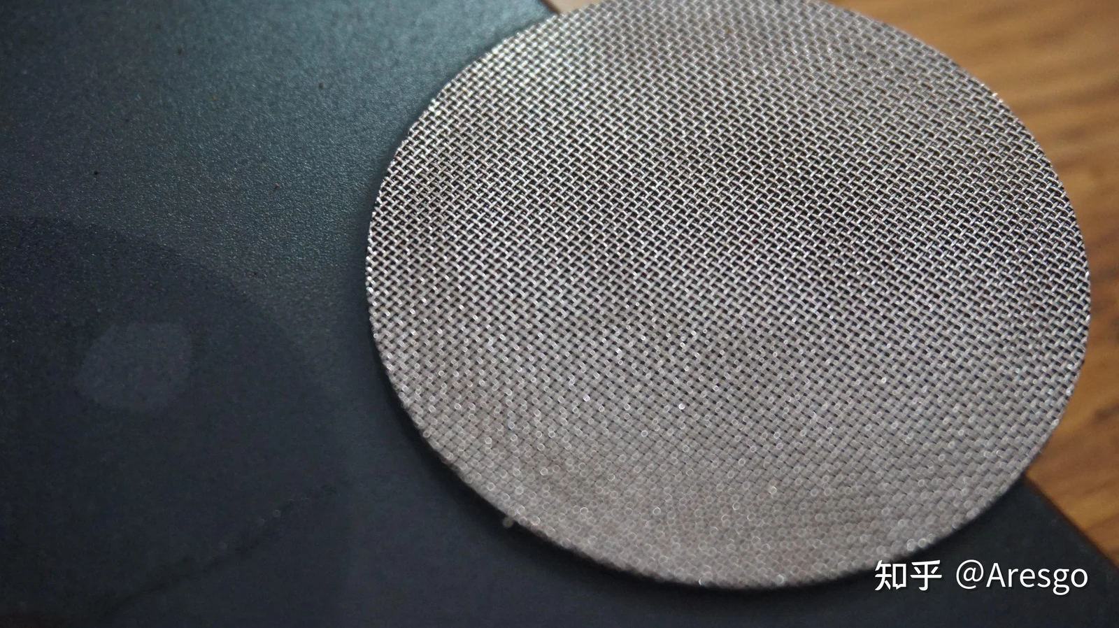 Close-up view of a circular stainless steel espresso puck screen resting on a dark surface.