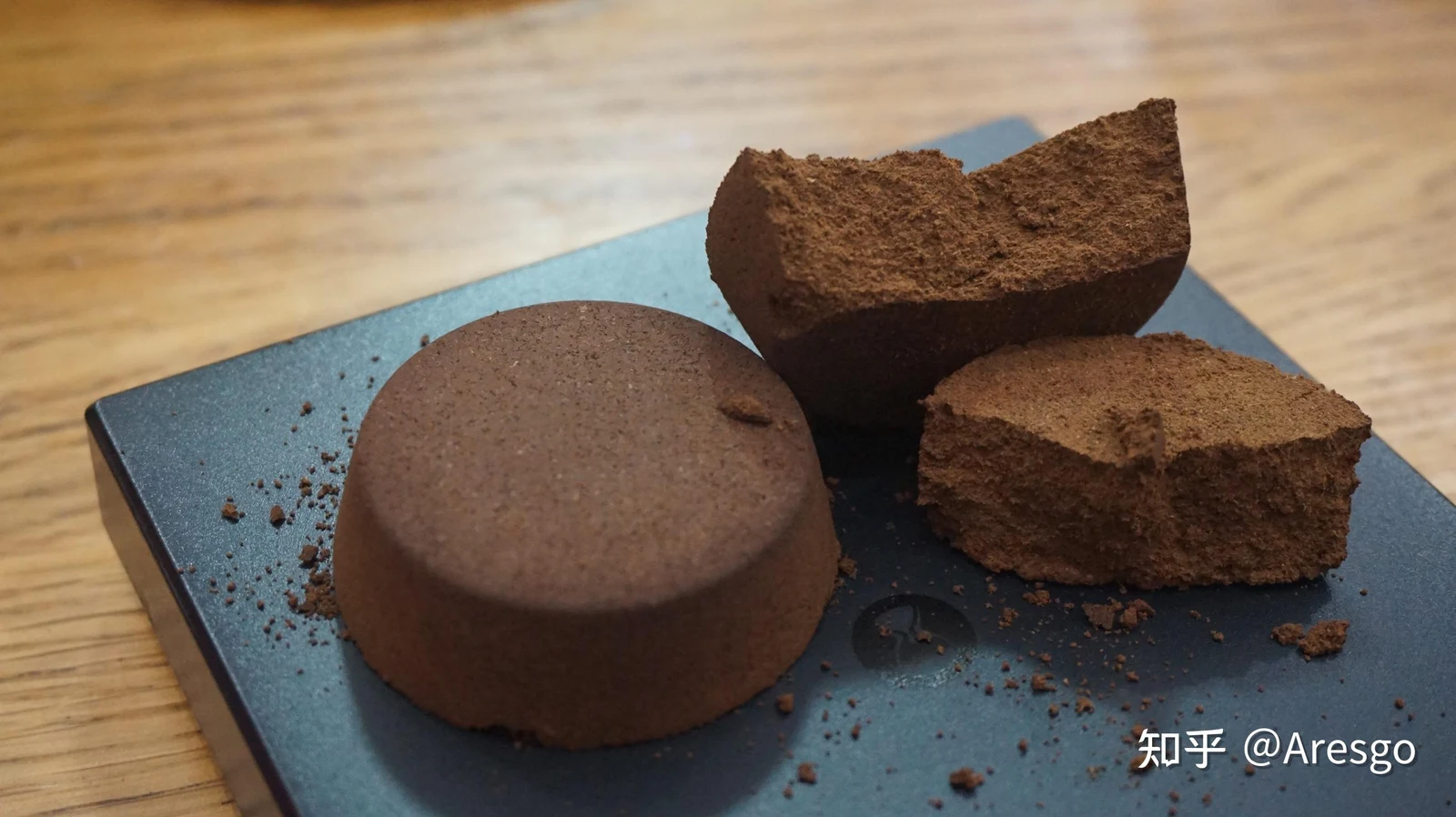 Extracted espresso coffee pucks, one intact and one broken in two pieces, displayed on a square dark platform atop a wooden table.