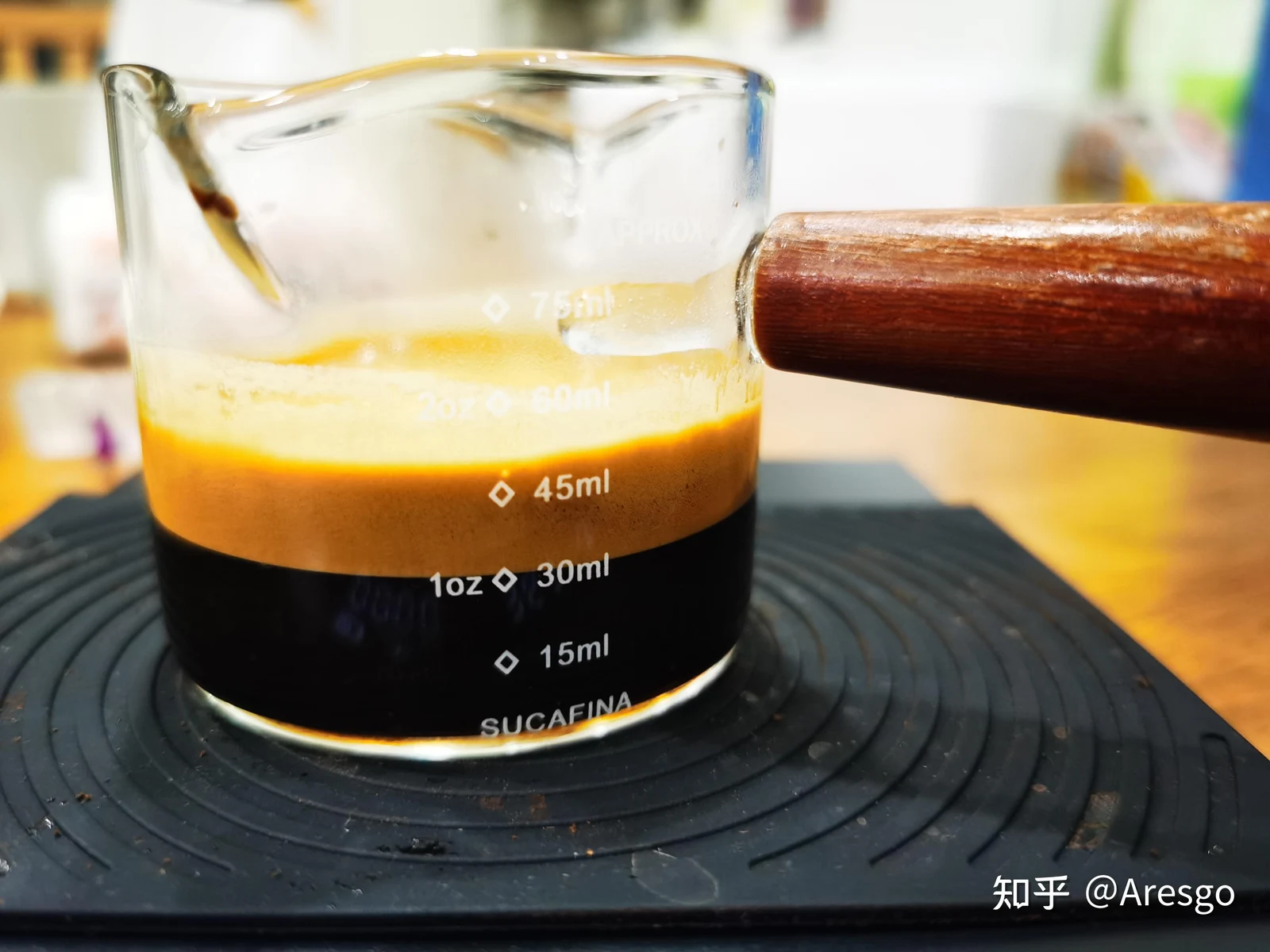 Clear glass espresso measuring cup with a wooden handle filled with freshly extracted espresso and thick crema, placed on a black scale mat.