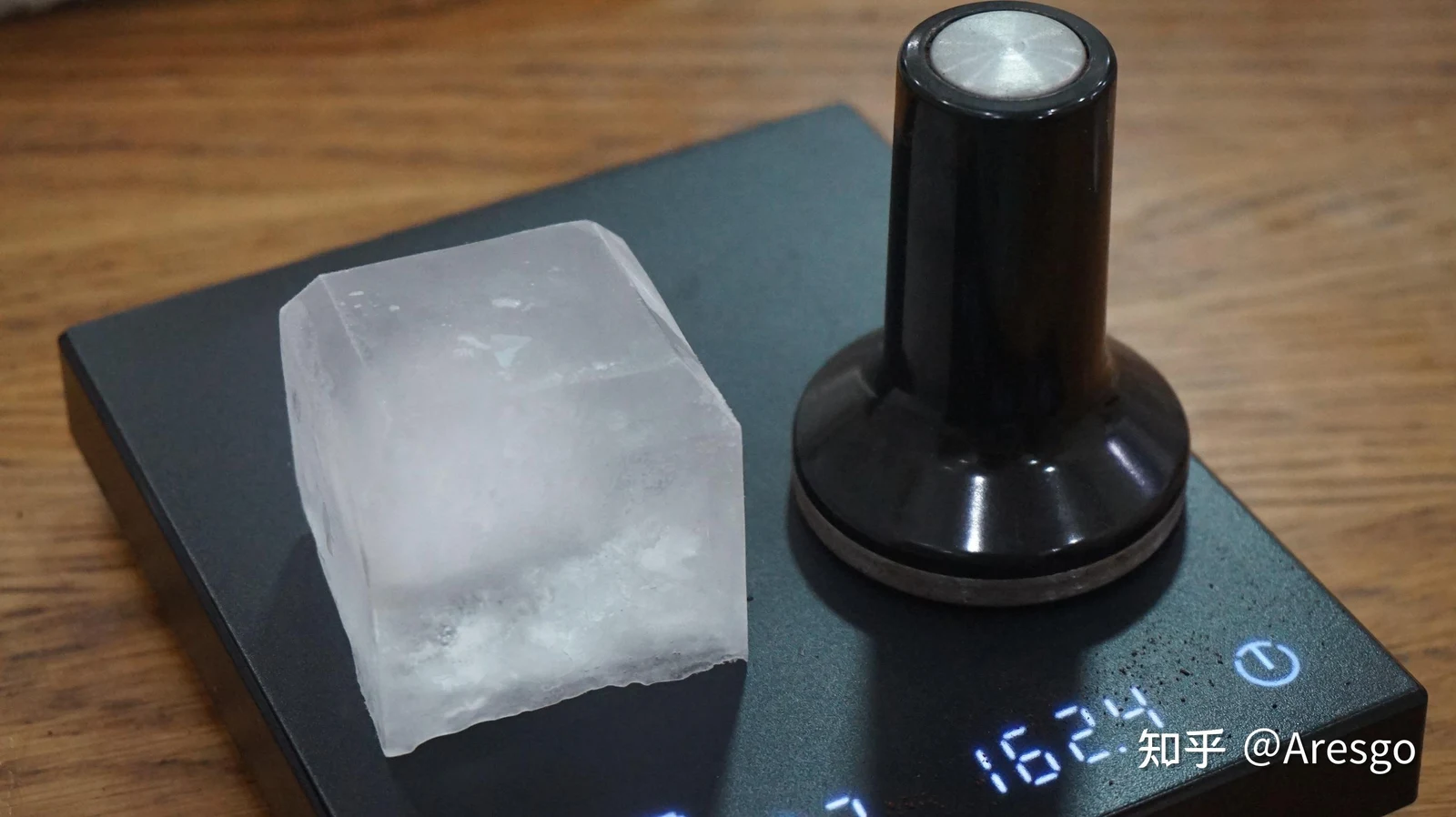 A large cubic ice cube and a black espresso tamper placed side by side on a digital coffee scale.