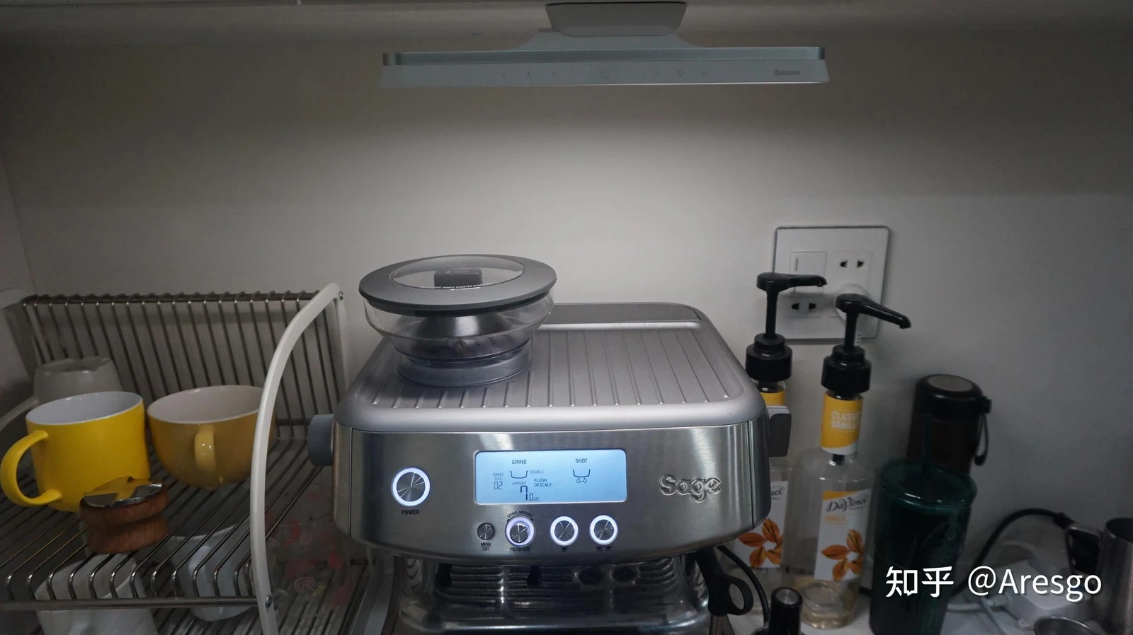 A stainless steel Sage 878 espresso machine illuminated by a slim light bar mounted underneath an upper cabinet, set beside syrup bottles and cups.