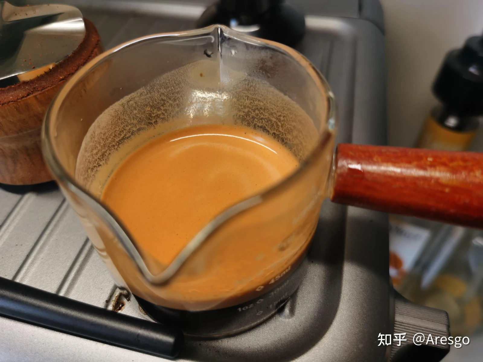 Clear glass espresso measuring pitcher with a wooden handle filled with freshly extracted espresso topped with thick golden crema, sitting on the drip tray of an espresso machine.