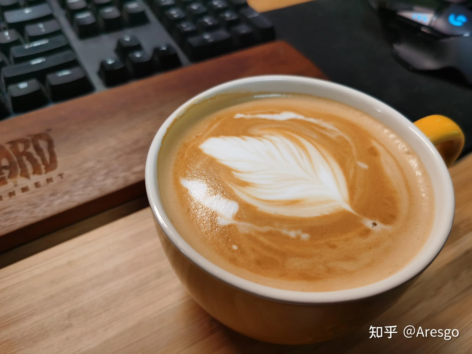 A cup of latte coffee with white leaf latte art on top, resting on a desk next to a wooden keyboard wrist rest and a mechanical keyboard.