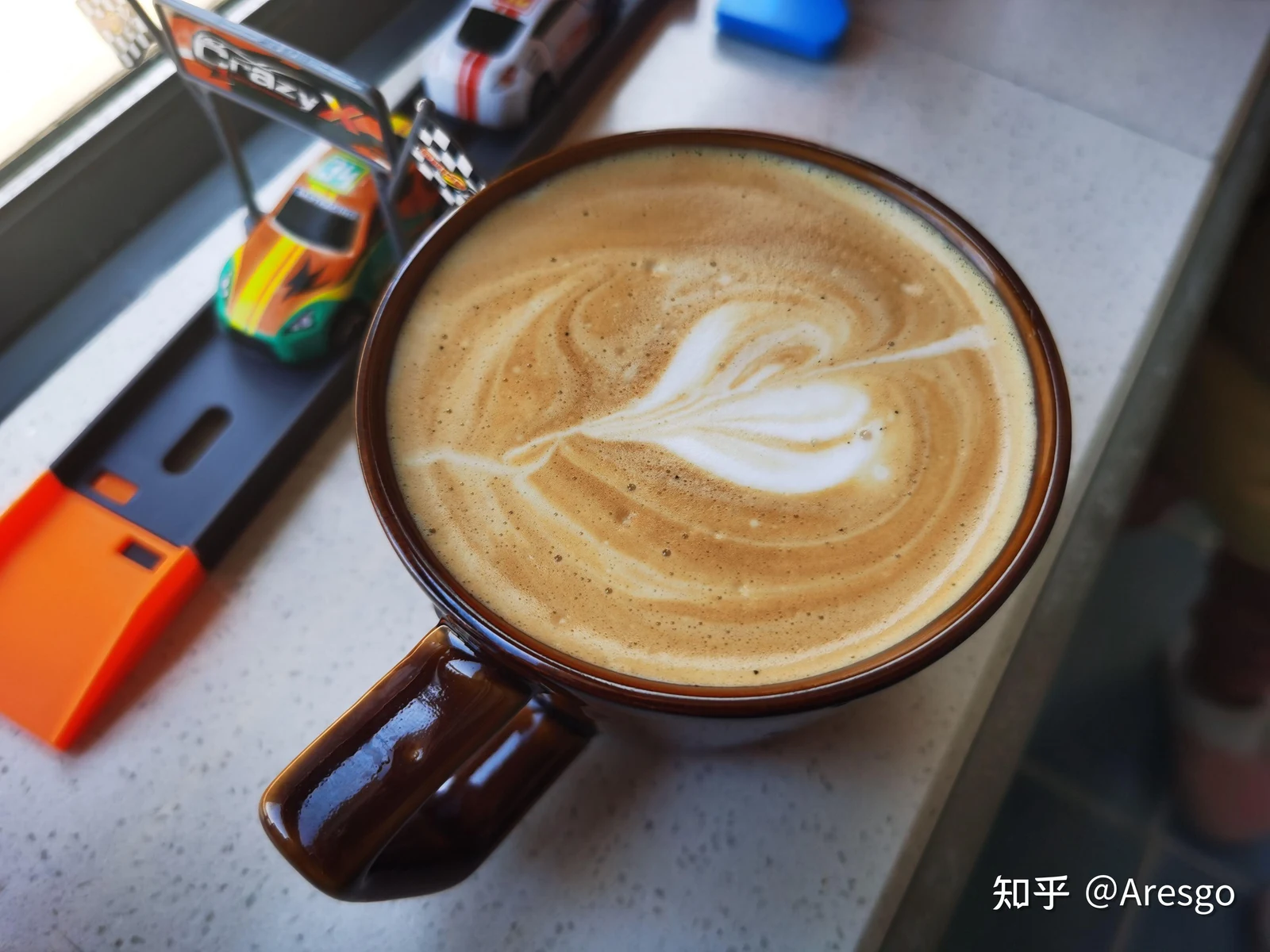 Top-down view of a brown ceramic mug filled with latte coffee featuring heart-shaped latte art, set on a windowsill next to a toy car track.