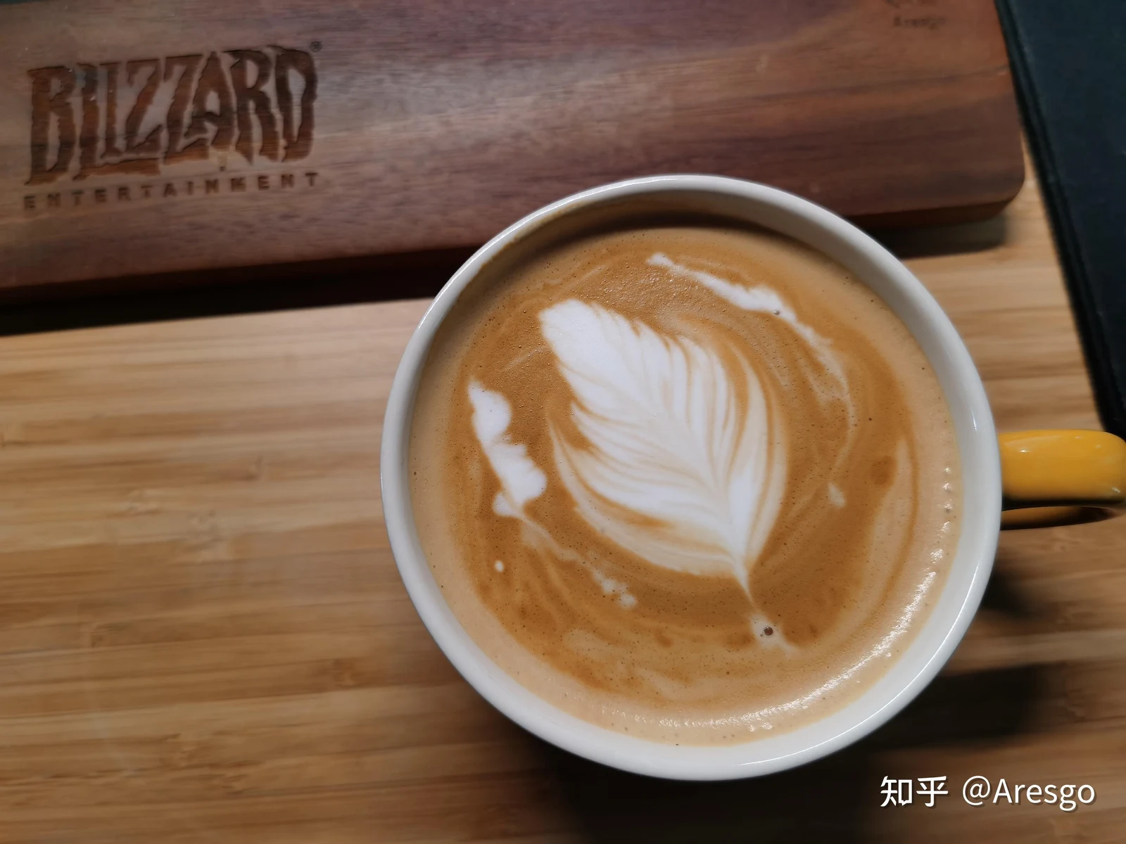Top-down view of a cup of coffee with white latte art in a leaf pattern, placed on a wooden surface next to a Blizzard Entertainment wooden board