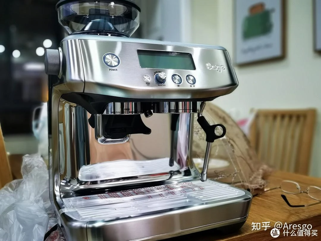 Stainless-steel Sage espresso machine with built-in grinder, front control panel, portafilter, steam wand, and drip tray on a wooden table.