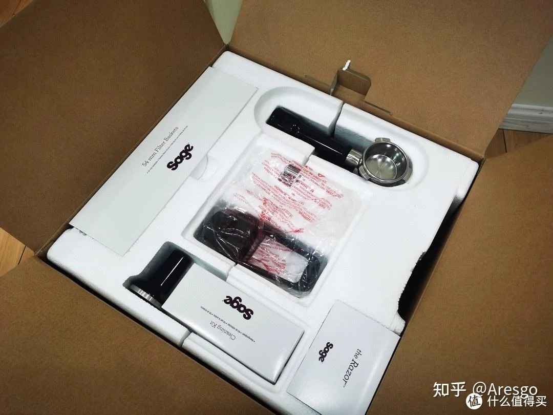 Open cardboard box containing a Sage espresso machine and accessories, including a portafilter, black handle, tamper, and manuals arranged in white foam packaging.