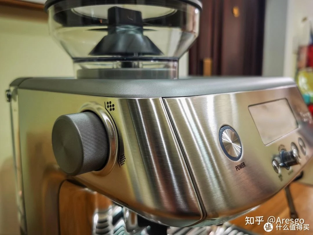 Close-up of a stainless-steel Sage 878 coffee machine with a bean hopper, a large grind-adjustment dial on the left, and a front control panel with a display and power button.