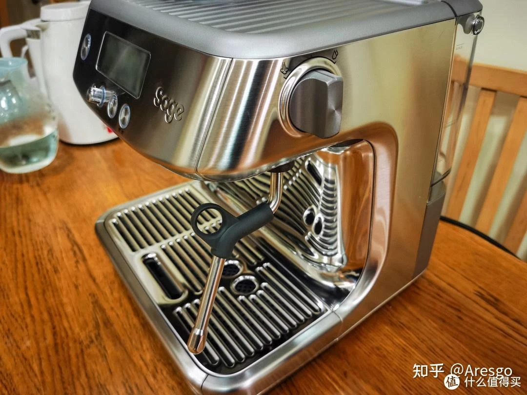 Stainless-steel Sage espresso machine on a wooden table, with a dark display and control buttons on the front, a large right-side dial, and a steam wand over the drip tray.
