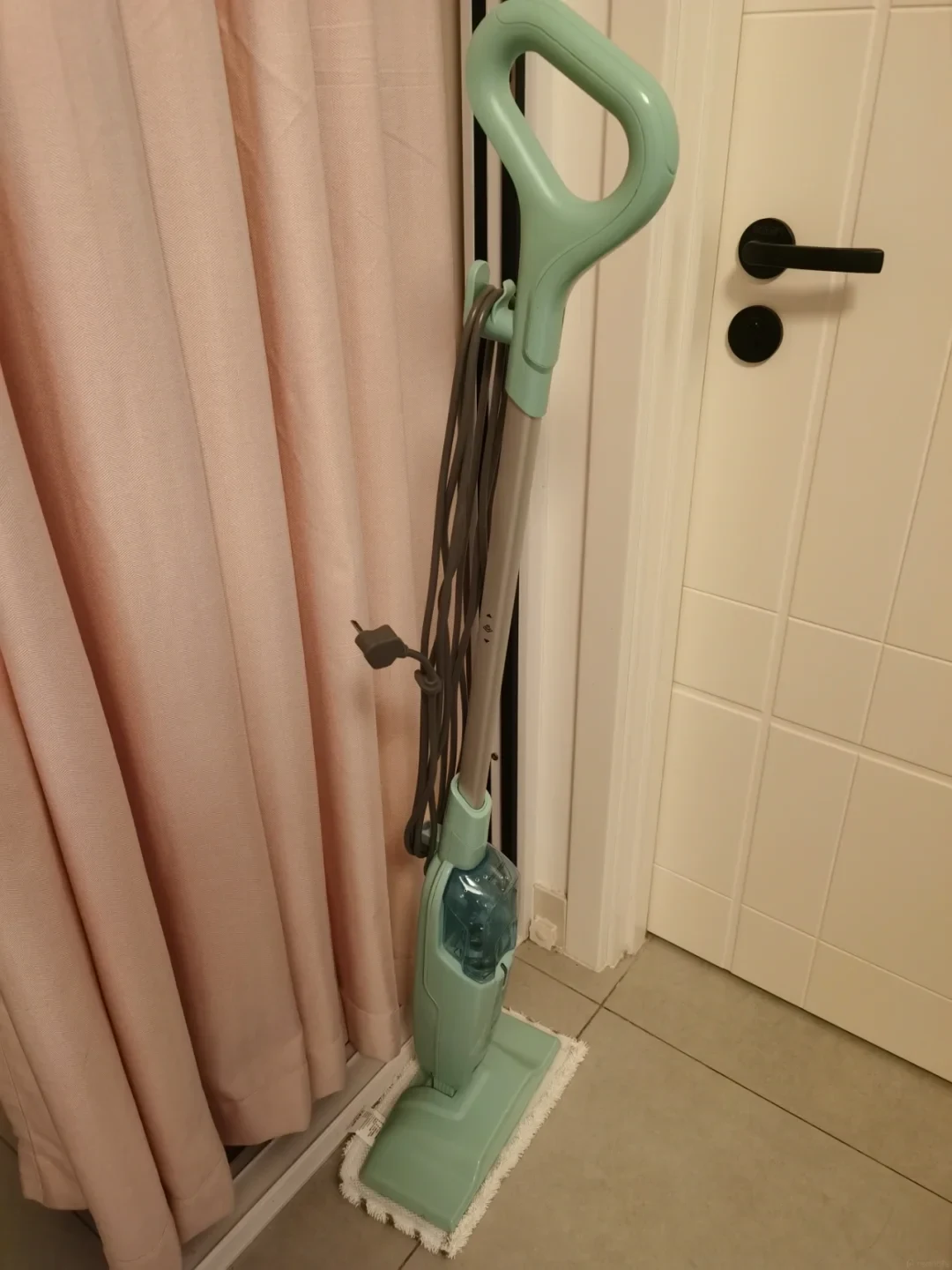 Mint green Shark steam mop standing upright on a floor cleaning pad between pink curtains and an interior door.