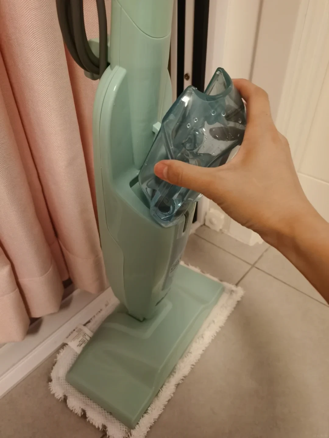 A hand inserts the removable transparent water tank into a light mint-green upright steam mop resting on a white mop pad.