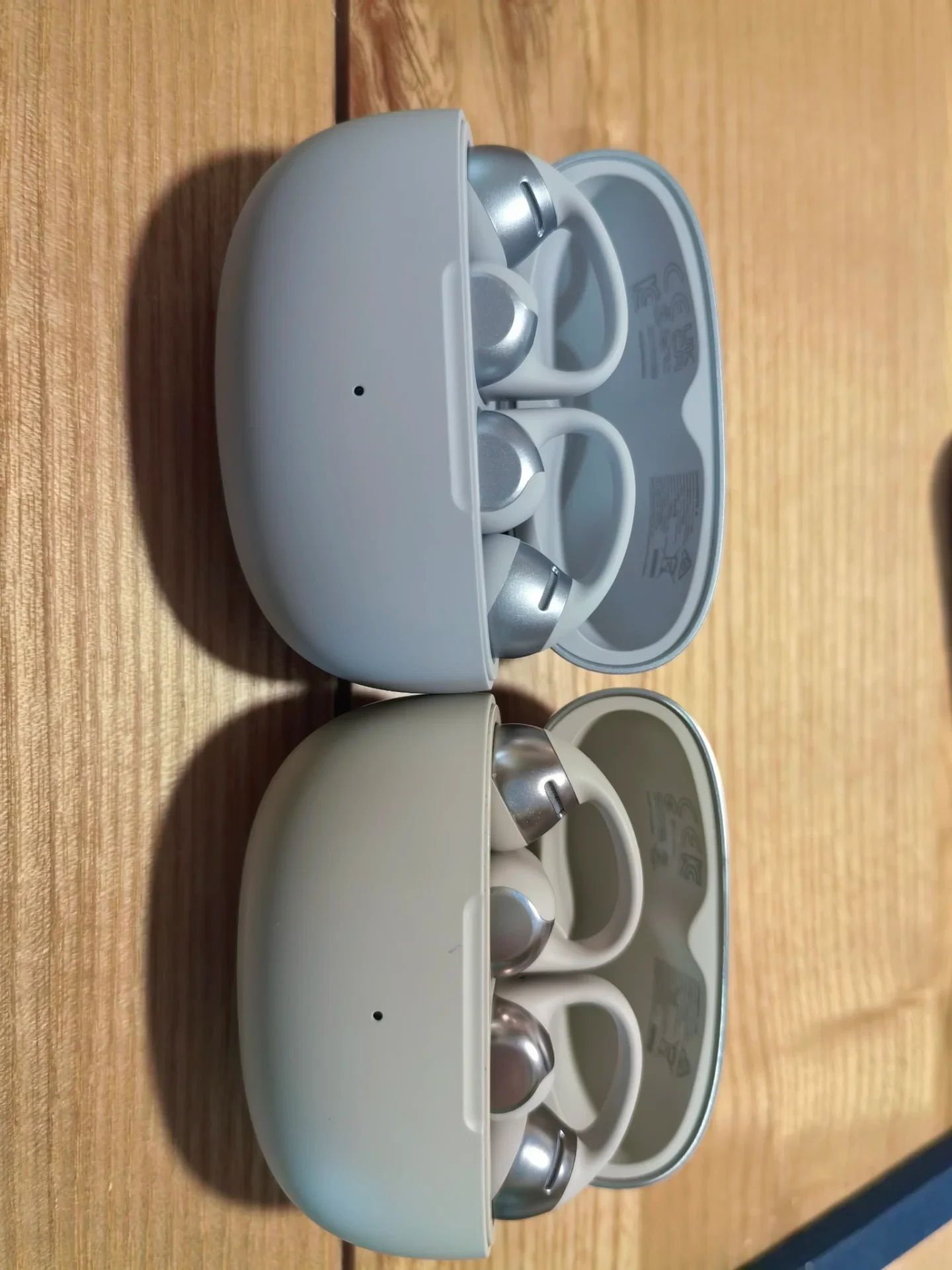 Two open Shokz earbud charging cases resting side by side on a wooden tabletop, displaying the curved clip-on open-ear earbuds seated inside.
