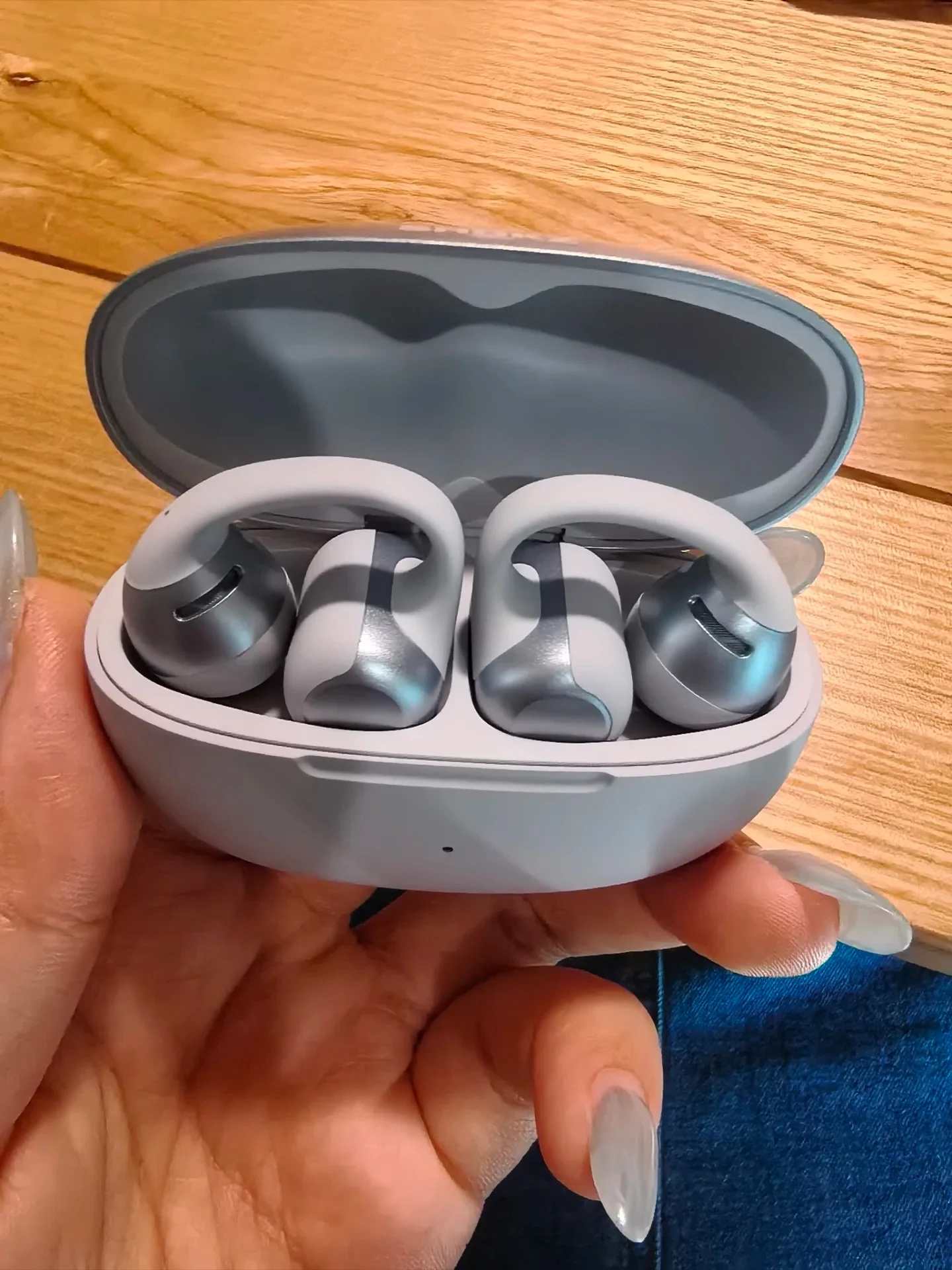 A hand holding an open light-gray charging case containing two clip-on open-ear earbuds with curved connectors and silver-accented driver modules.