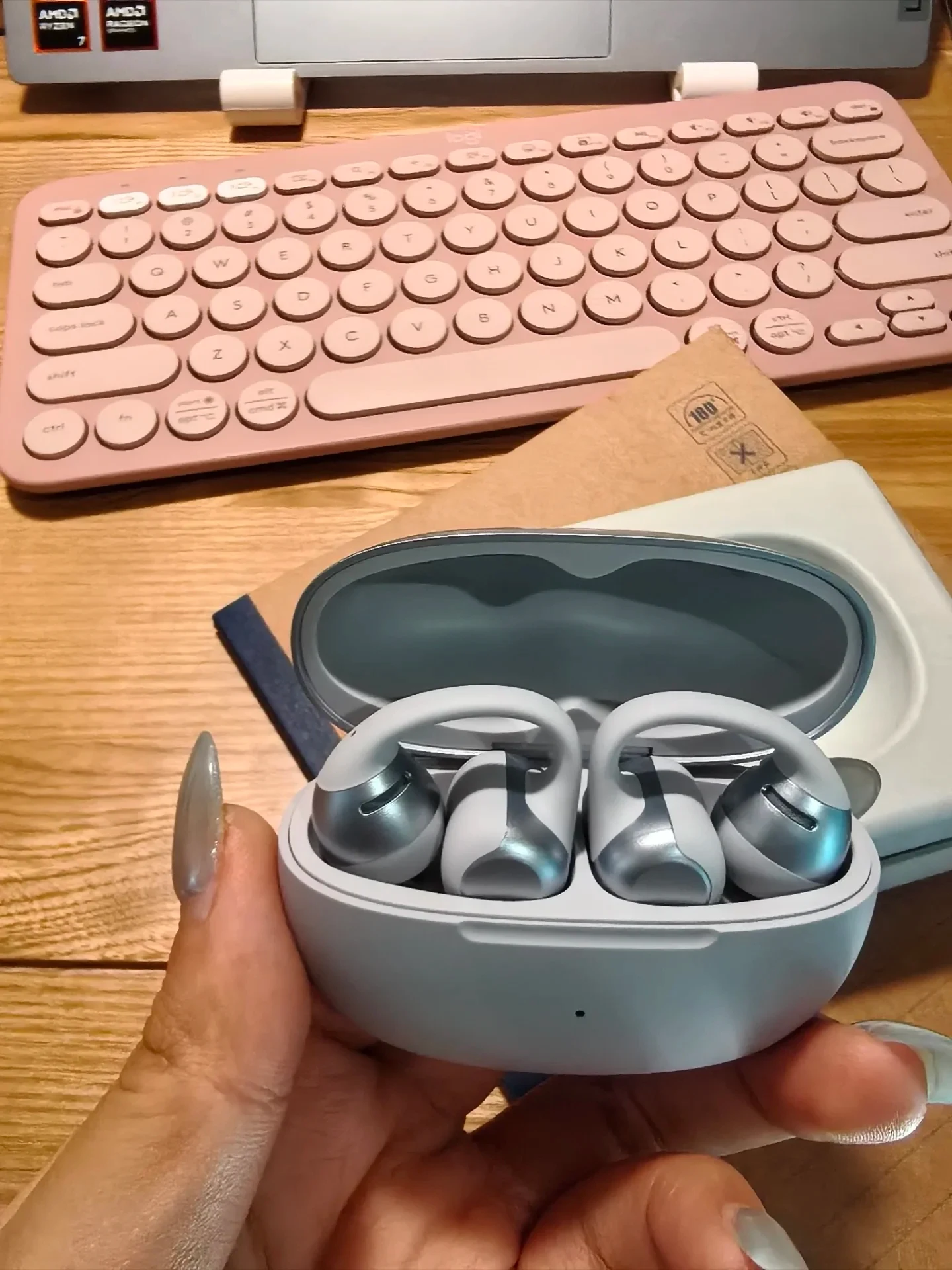 A person's hand holding open an oval charging case containing a pair of light gray and silver clip-on open-ear earbuds above a wooden desk with a pink keyboard.