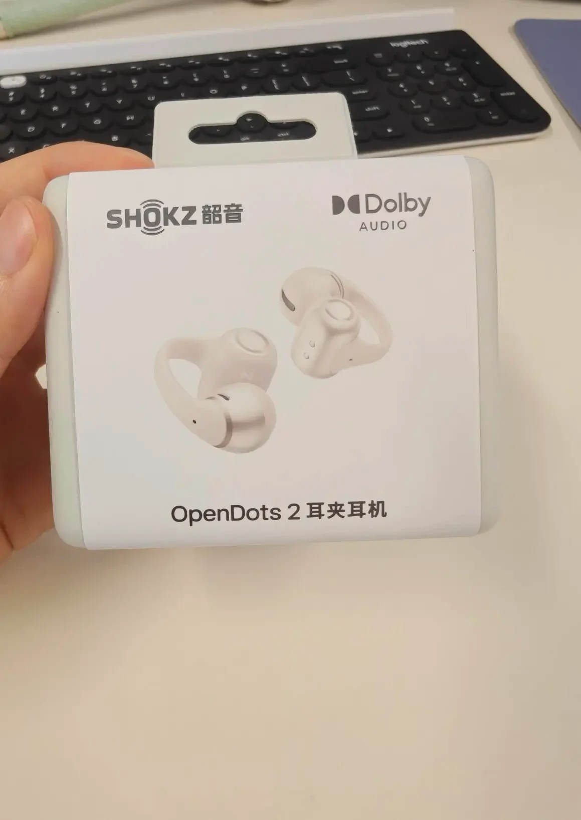 A person holding the retail box of Shokz OpenDots 2 clip-on earbuds above an office desk with a keyboard.