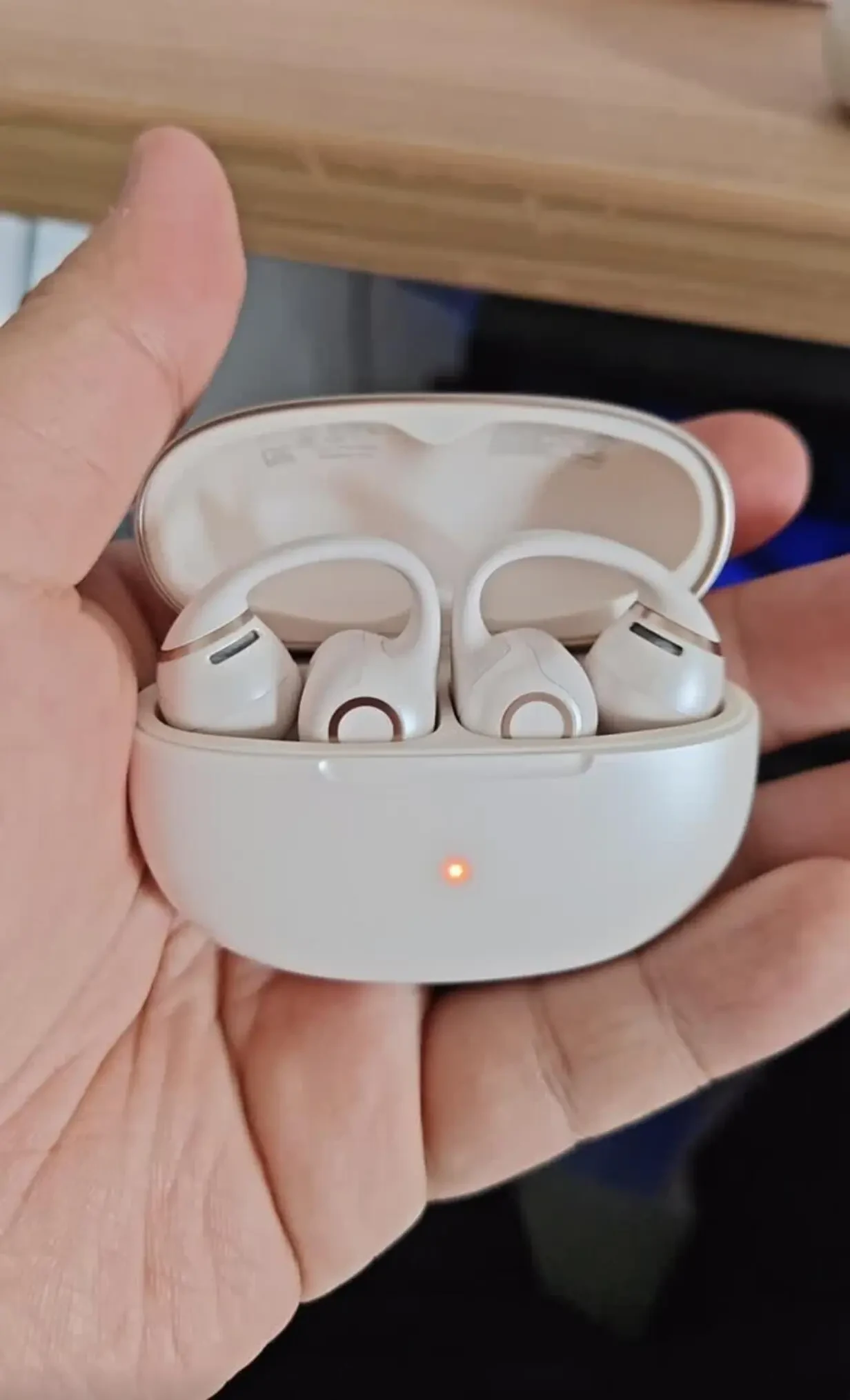 A person holding an open white charging case revealing a pair of beige clip-on open-ear earbuds resting inside, with an illuminated orange indicator light on the front of the case.