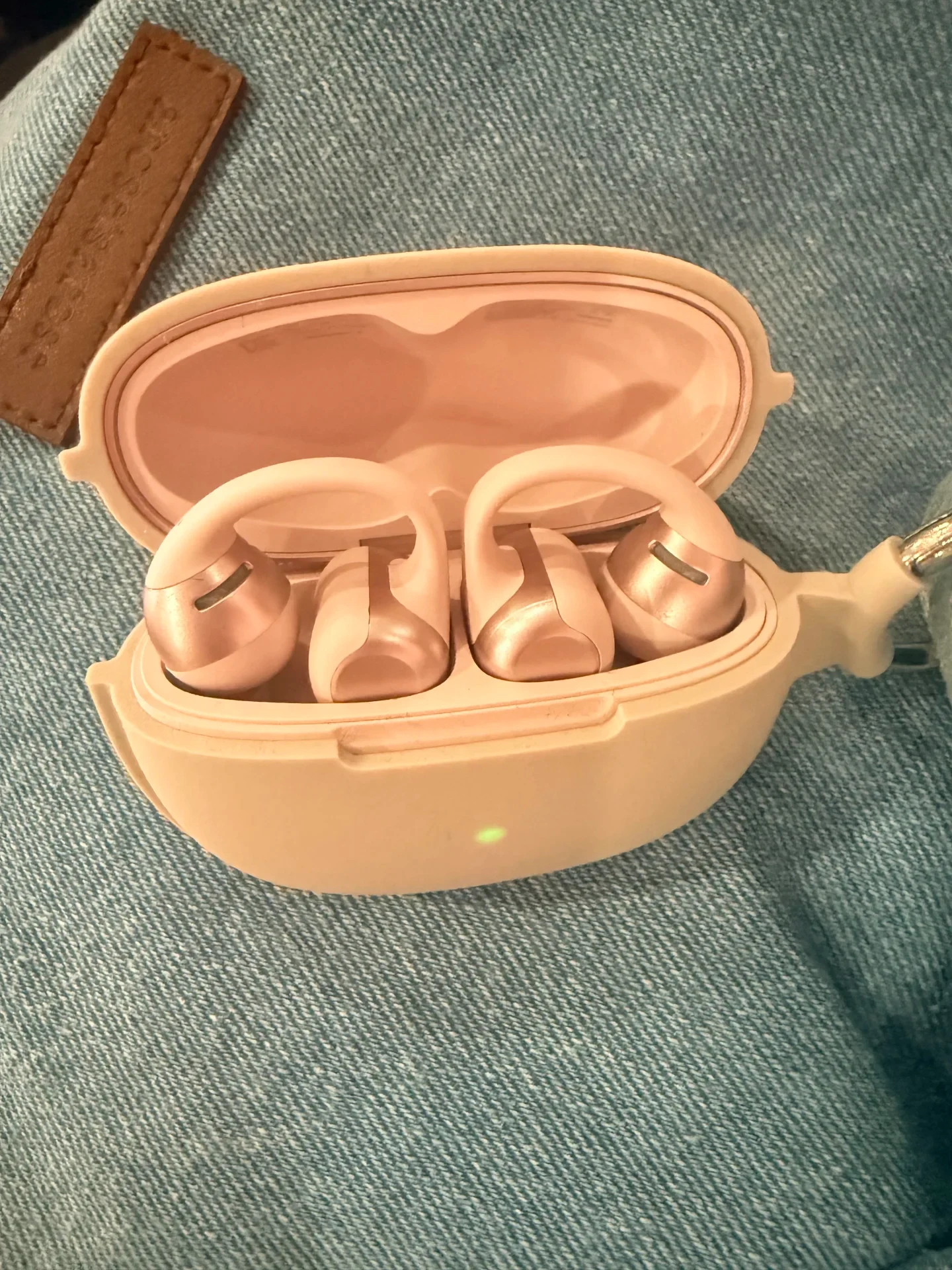 Pale pink open-ear clip earbuds resting in an open charging case with a protective silicone cover and a green indicator light, on blue denim fabric.