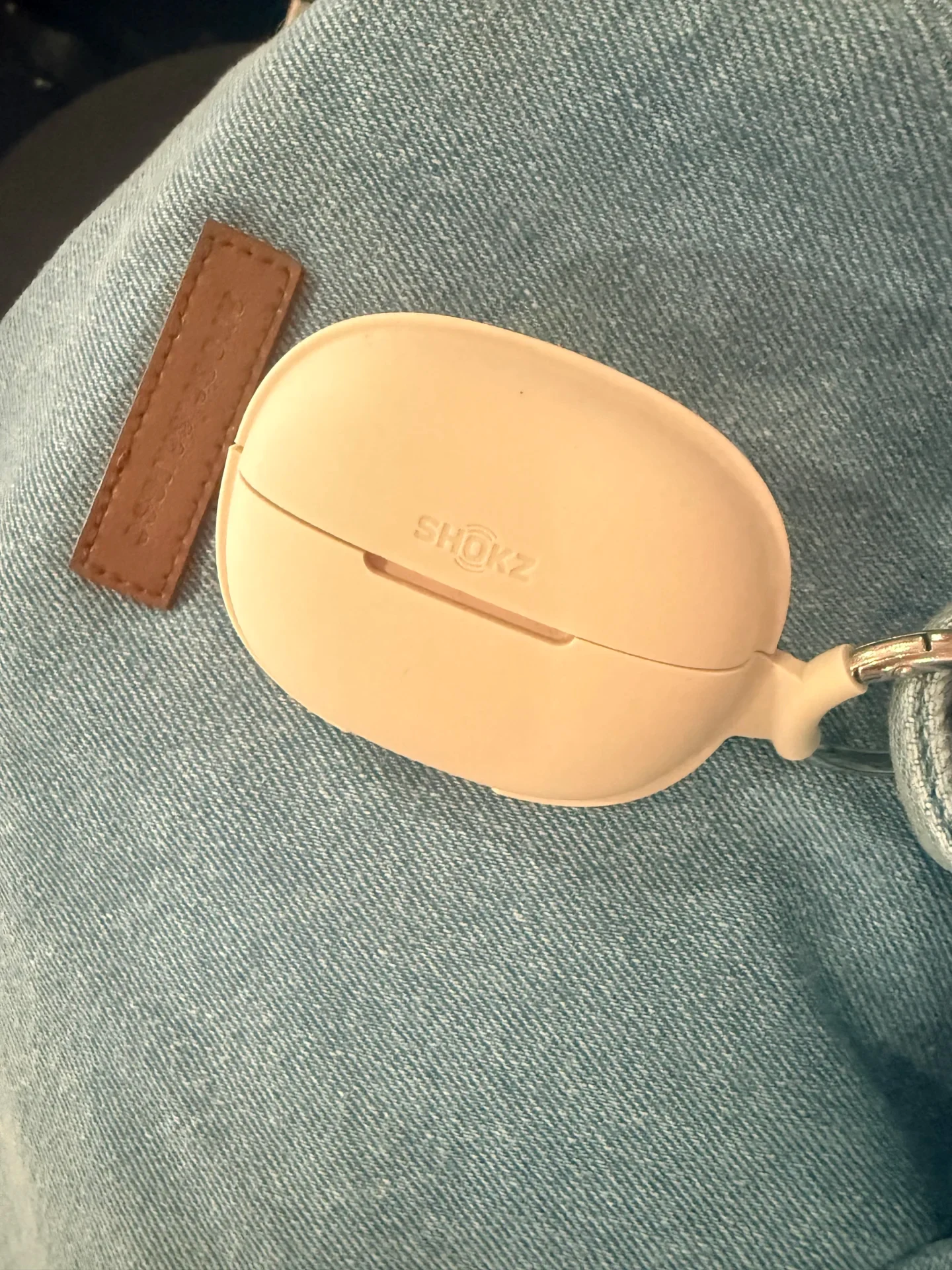 A closed beige Shokz earbud charging case in a silicone protective sleeve rests on light blue denim beside a brown stitched leather tag and metal clip.