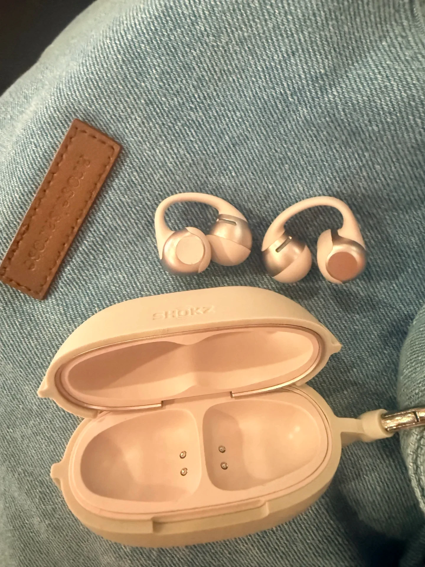 A pair of beige Shokz OpenDots 2 clip-on open-ear earbuds resting above their open charging case on light blue denim fabric.