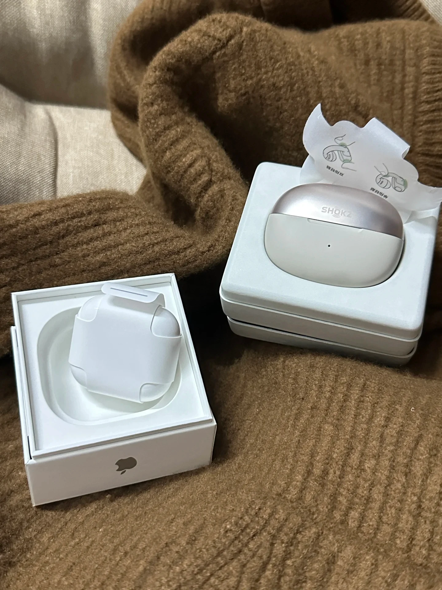 Two earphone boxes set side by side on a brown knit sweater, showing an Apple AirPods charging case wrapped in paper in its packaging tray on the left and a Shokz OpenDots ONE charging case in its tray on the right with an instructional paper tab.