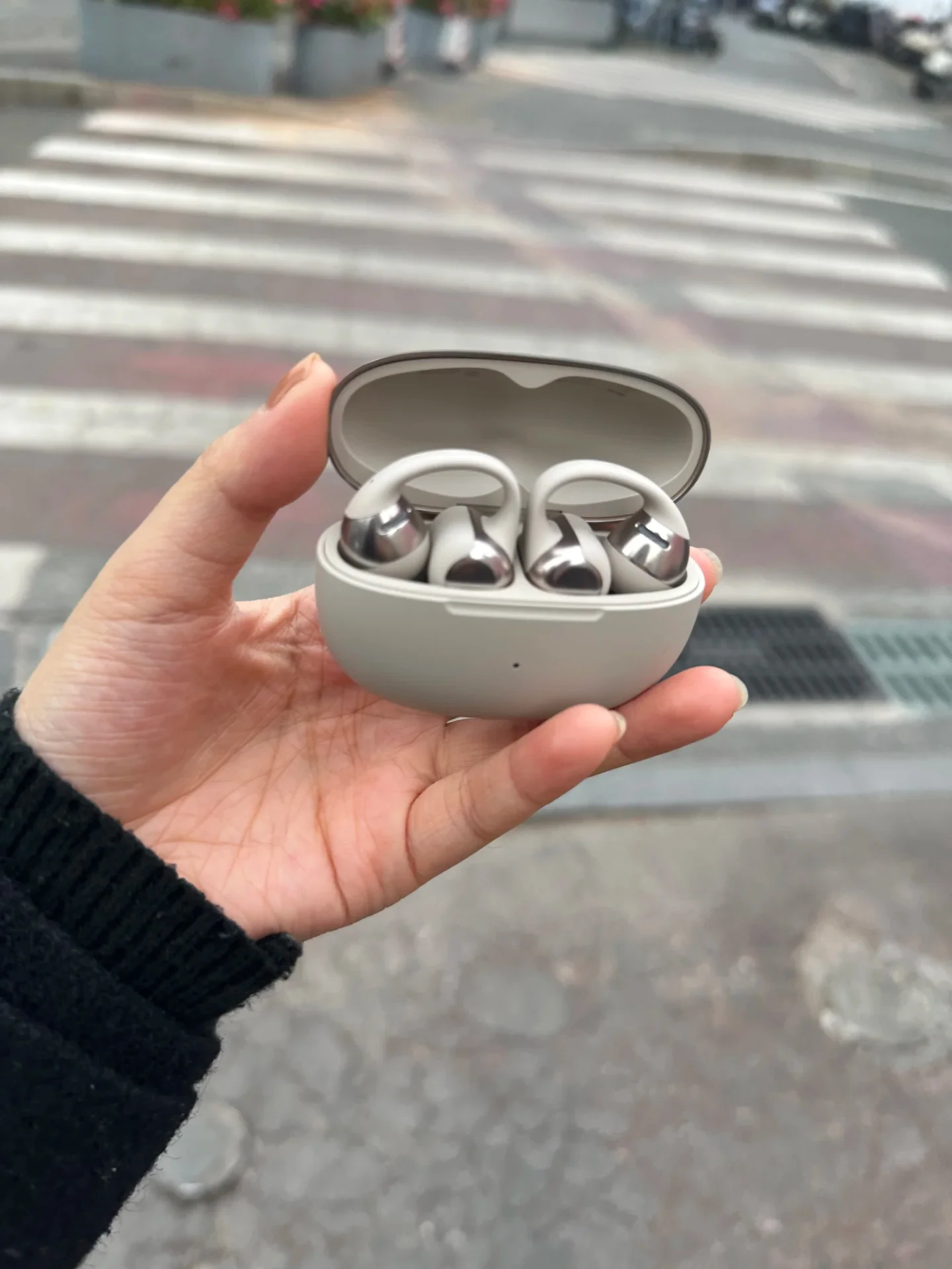 A hand holds open a light grey charging case outdoors to display two Shokz OpenDots ONE clip-on earbuds resting inside against a blurred street backdrop.
