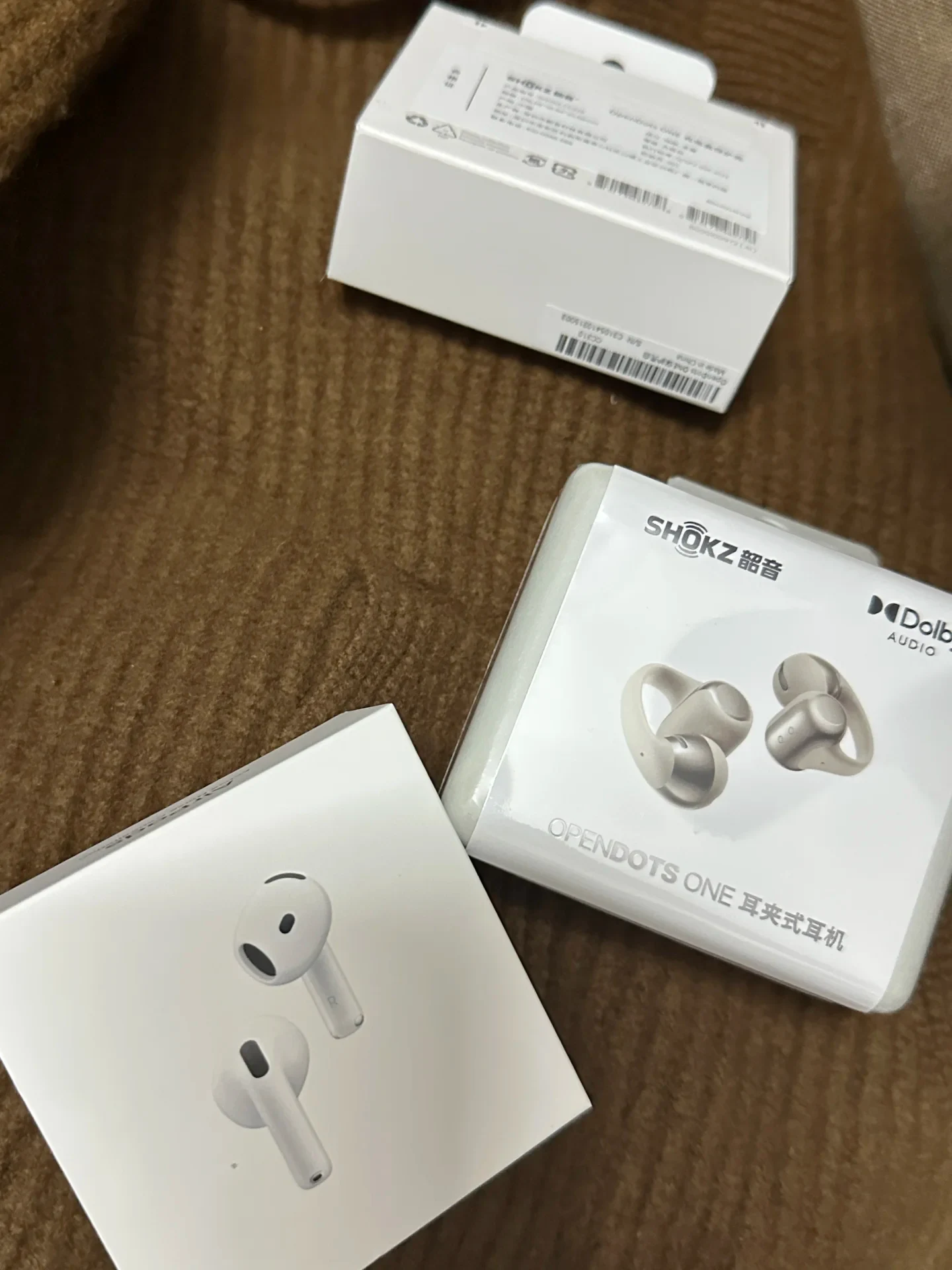 Retail packaging for Shokz OpenDots ONE clip-on earbuds and Apple AirPods resting on a brown ribbed knit fabric.