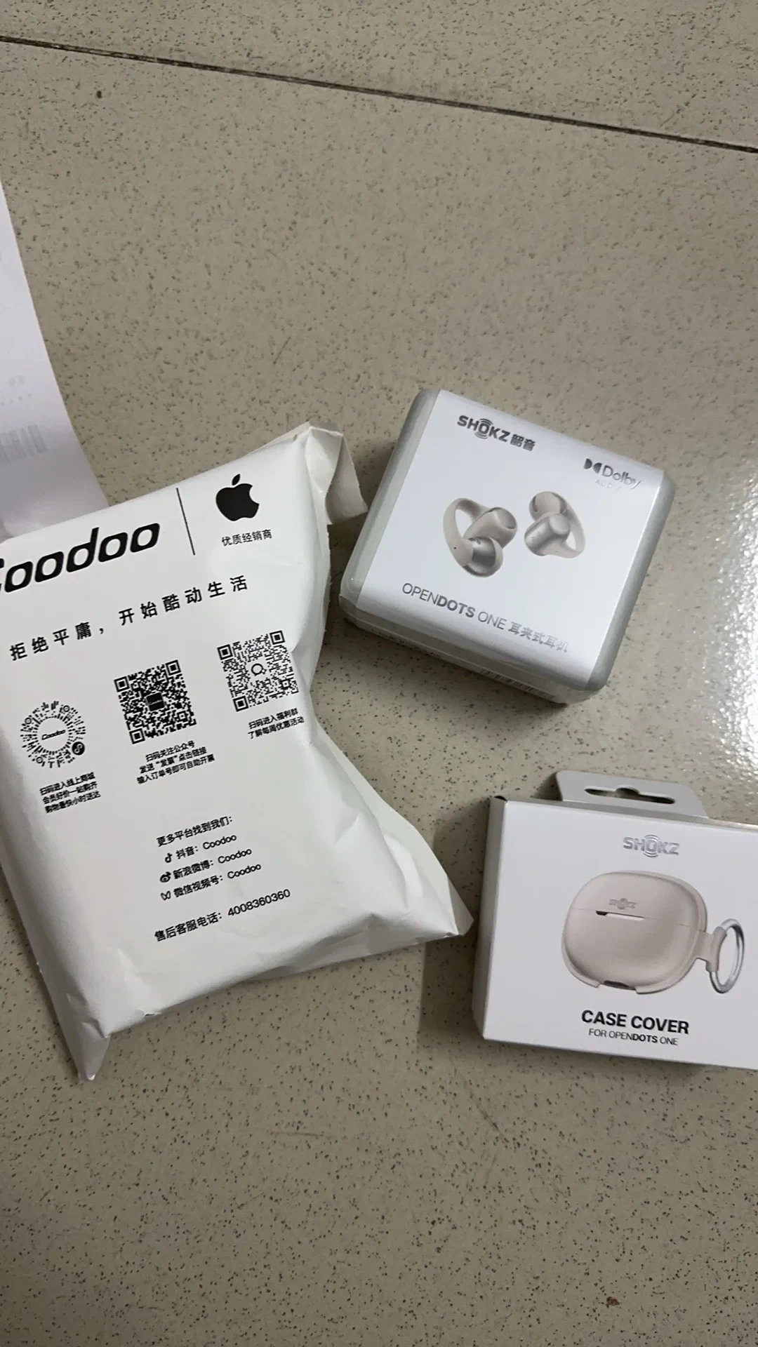 A packaged pair of Shokz OpenDots ONE clip-on earphones in their retail box, alongside a separate box for an OpenDots ONE case cover and a white Coodoo retailer shipping pouch on a tiled floor.