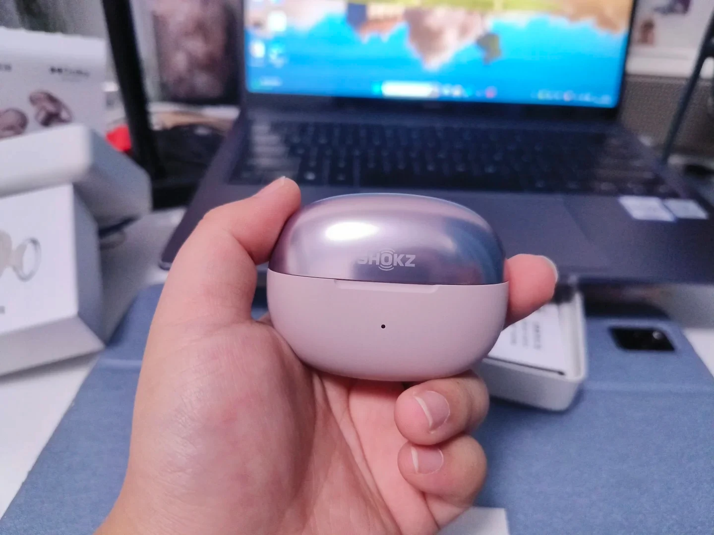 A person's hand holding the closed compact charging case of Shokz OpenDots ONE earbuds, featuring a metallic lilac lid with the Shokz logo and a matte light pink lower body, with a laptop and open packaging in the blurred background.