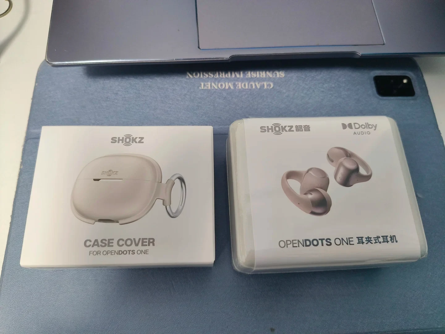 Two product packaging boxes on a blue desk mat beneath a laptop: a Shokz case cover box for OpenDots One on the left and a shrink-wrapped Shokz OpenDots One clip-on earphone box on the right.