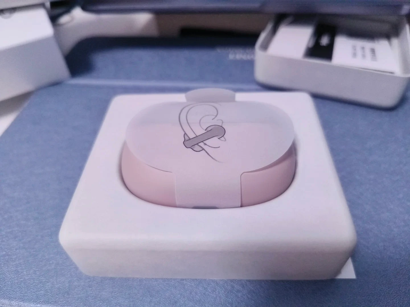 A light pink earbud charging case seated in a white molded packaging tray, wrapped in a translucent protective sleeve with a line illustration demonstrating how to clip the earbud onto an ear.