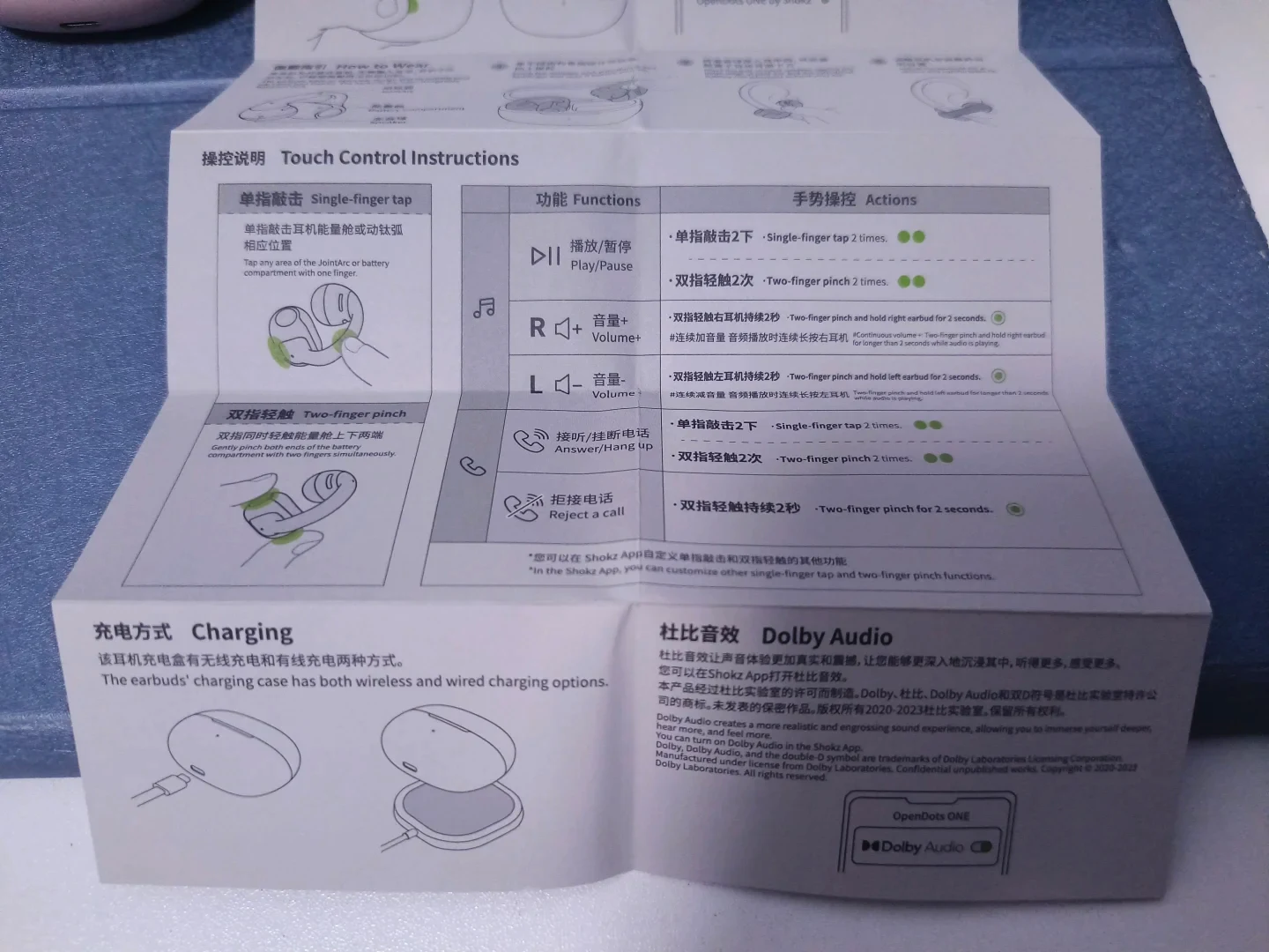 A printed bilingual user guide for the Shokz OpenDots ONE earphones, displaying touch control instructions, diagrams for single-finger tapping and two-finger pinching, charging methods, and Dolby Audio details.