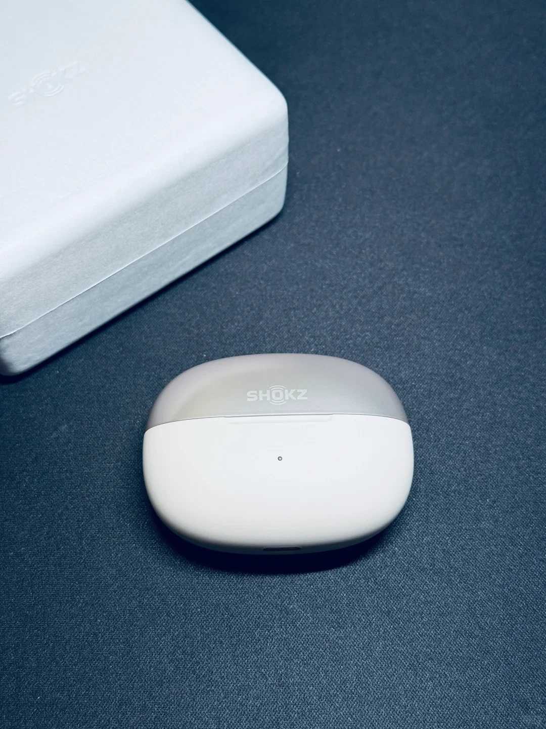 A closed Shokz wireless earbuds charging case resting on a dark textured surface, positioned below a white packaging box in the upper left corner.