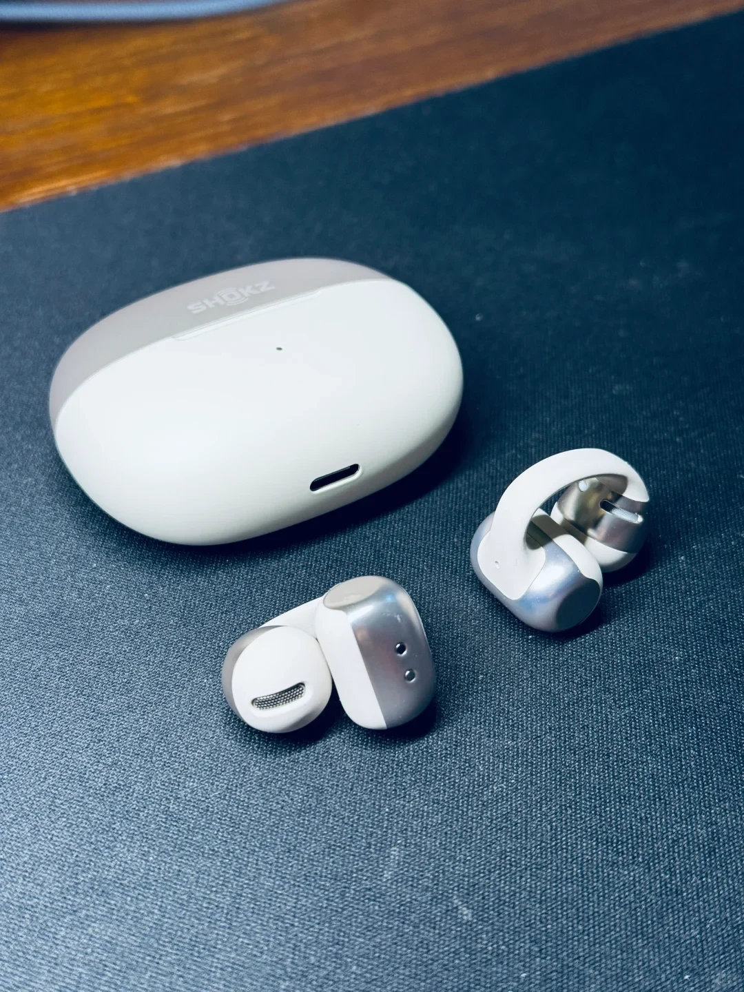 A pair of white and metallic silver clip-on open-ear earbuds shown beside their matching pebble-shaped charging case with the Shokz logo, placed on a dark textured desk mat.