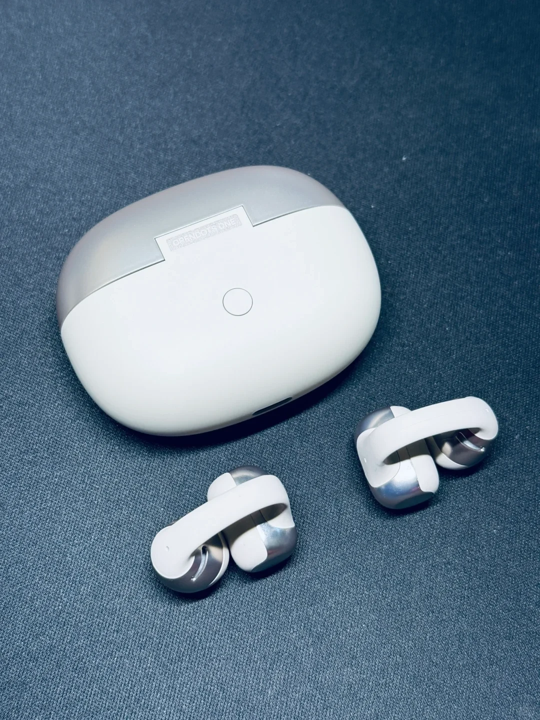 A white and metallic silver wireless earphone charging case resting on a dark textured background alongside a pair of open-ear clip-on earbuds.
