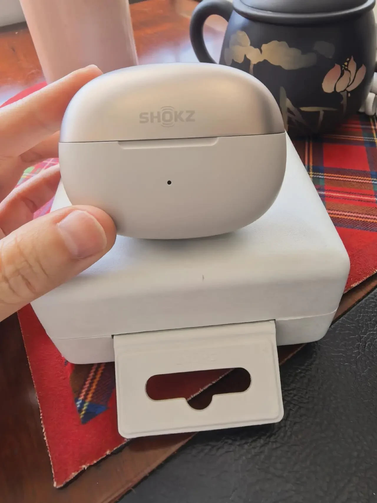A person's hand holding the closed two-tone charging case of Shokz clip-on earbuds directly above its white inner packaging box, which has a hanging tab attached.