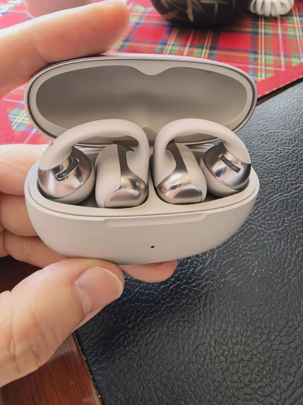 A close-up of a hand holding an open light-gray charging case containing two clip-on open-ear earbuds with metallic silver speaker capsules.