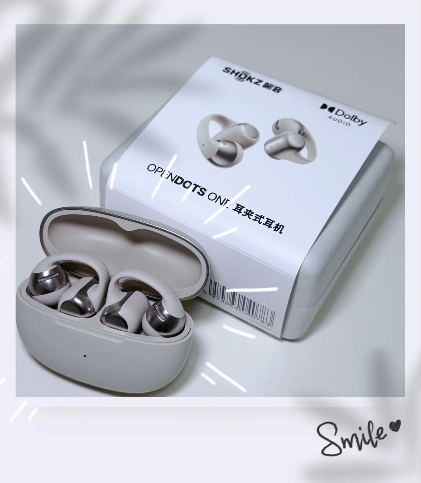 An open charging case containing Shokz OpenDots ONE clip-on earbuds resting beside its retail box on a light surface.