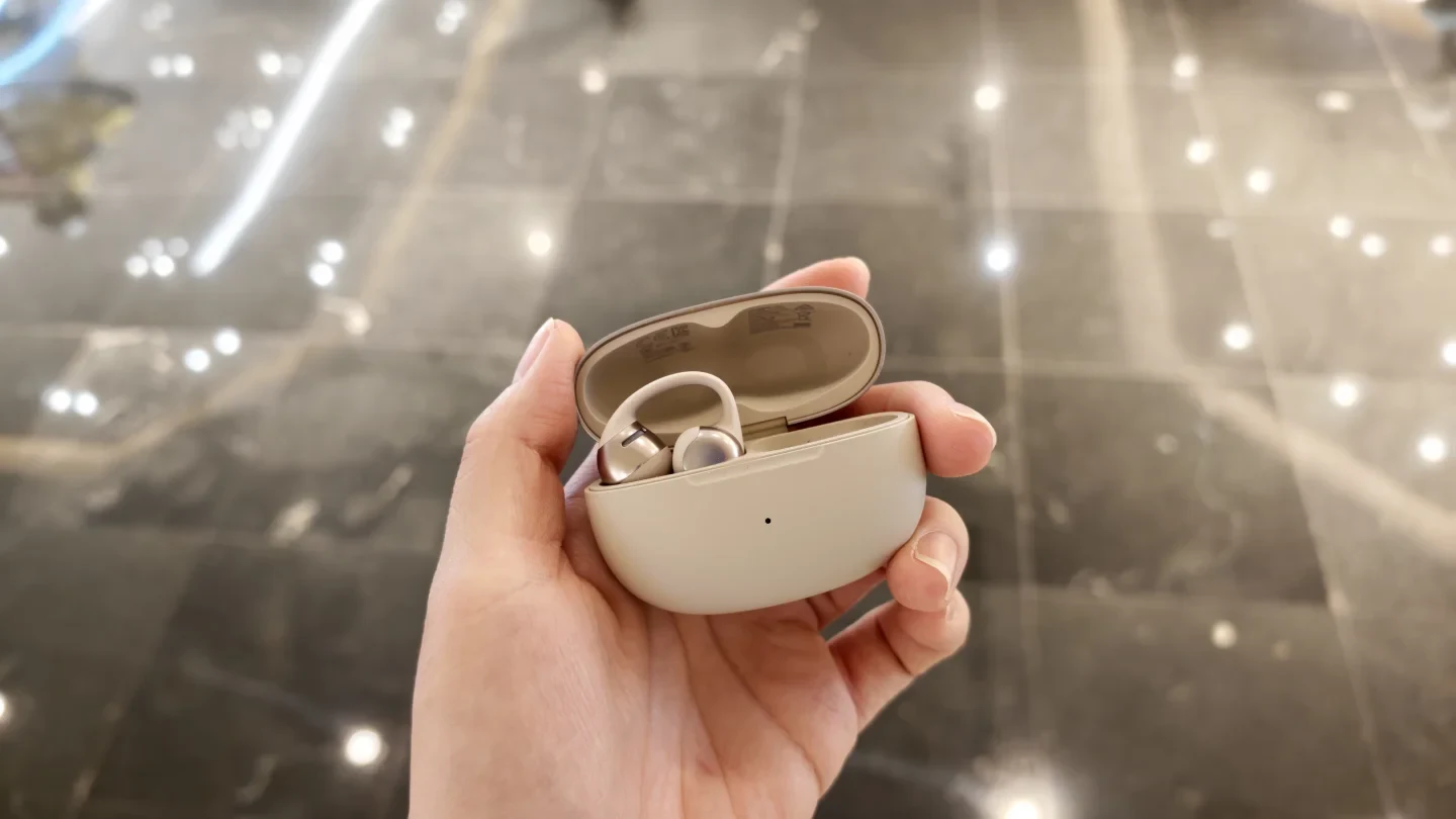 A person holds open a light-colored charging case for clip-on earbuds, showing one earbud resting inside while the adjacent slot is empty against a tiled floor background.