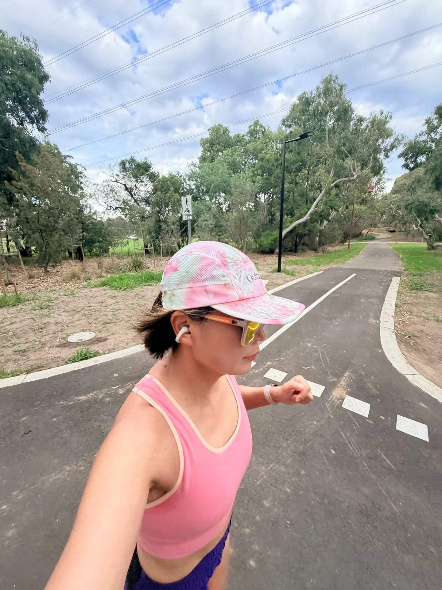 A runner wearing a pink top, sunglasses, a colorful cap, and a white clip-on earbud jogs along an outdoor paved path lined with trees.