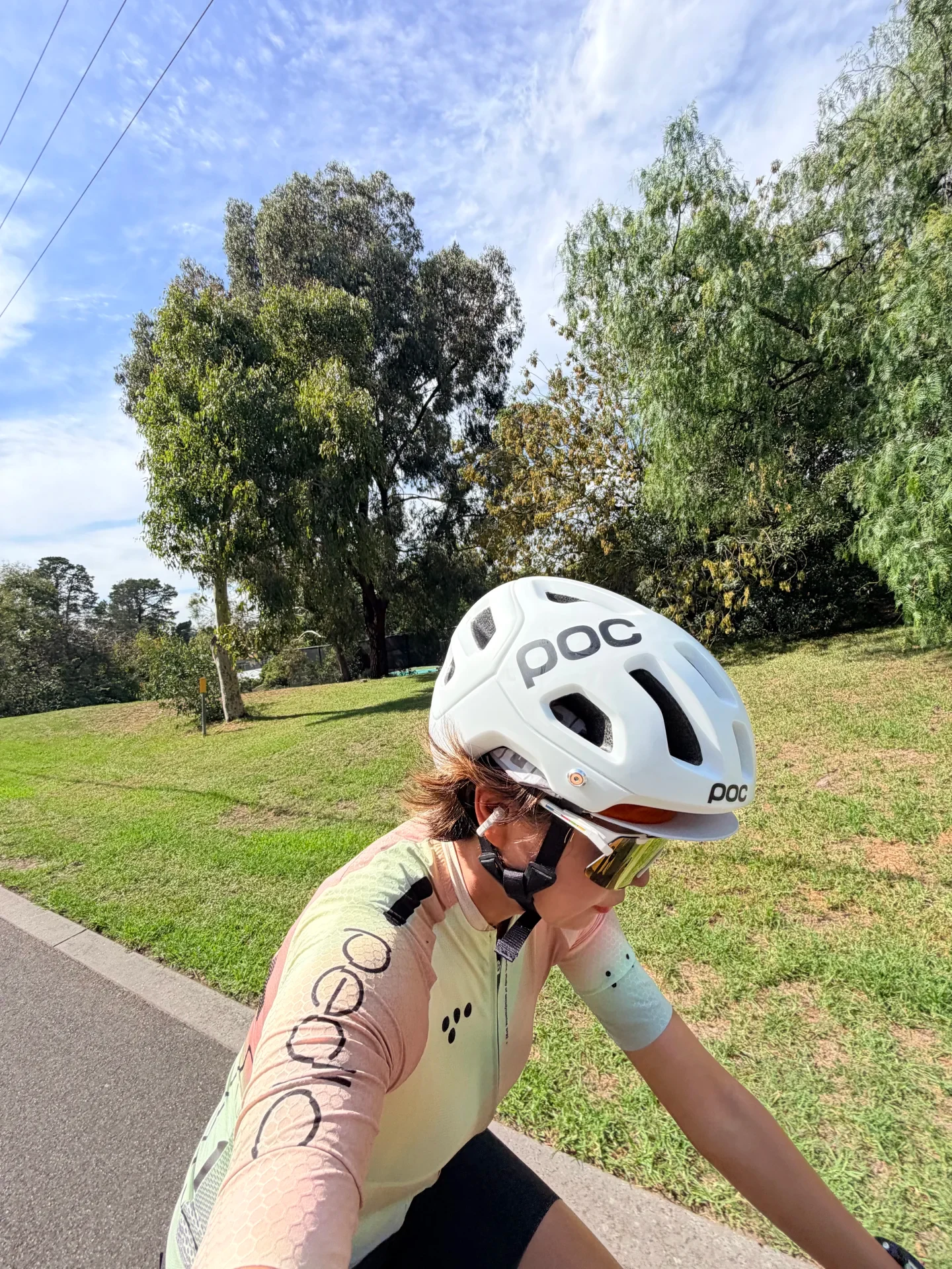 A cyclist wearing a white helmet, cycling sunglasses, and a long-sleeve jersey rides on a paved outdoor path with green trees in the background, with a white clip-on earbud attached to her right ear.